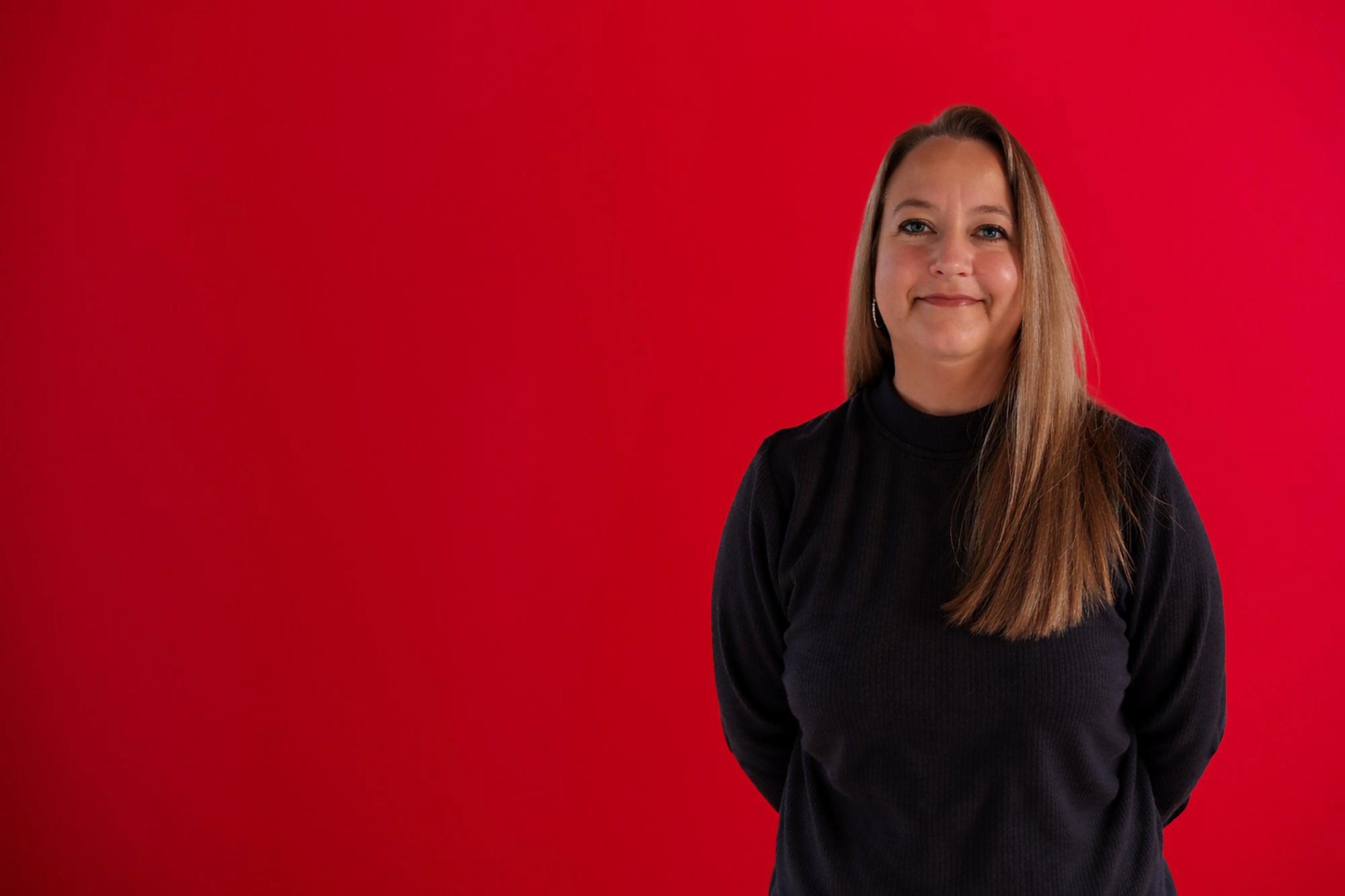 Portrait of a woman with long blonde hair wearing a white top and a long silver chain necklace, standing against a red background.
