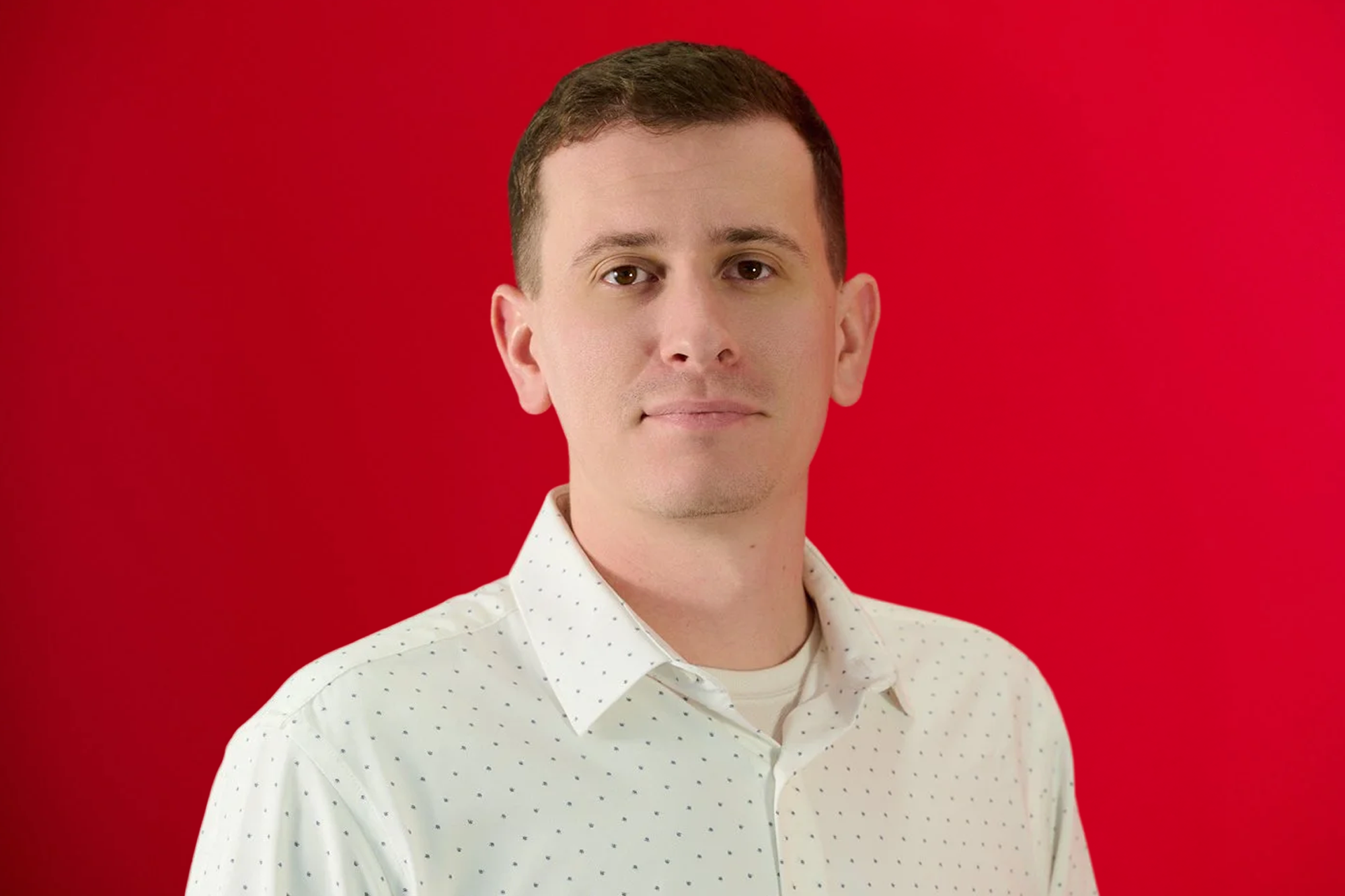 Portrait of a young man with short brown hair and light skin, wearing a white shirt with small dark polka dots, standing against a solid red background.