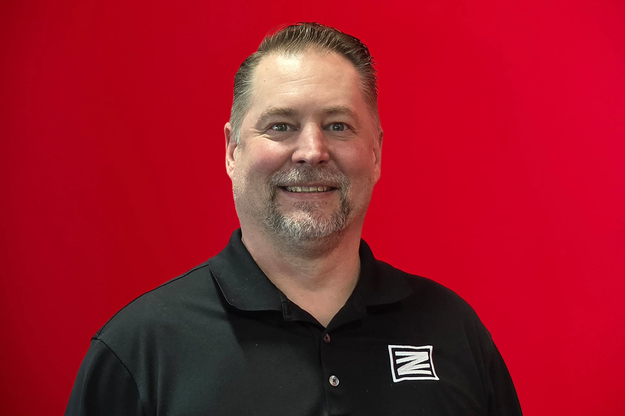 A man with a beard, wearing a gray polo shirt with a logo, smiling in front of a red background.