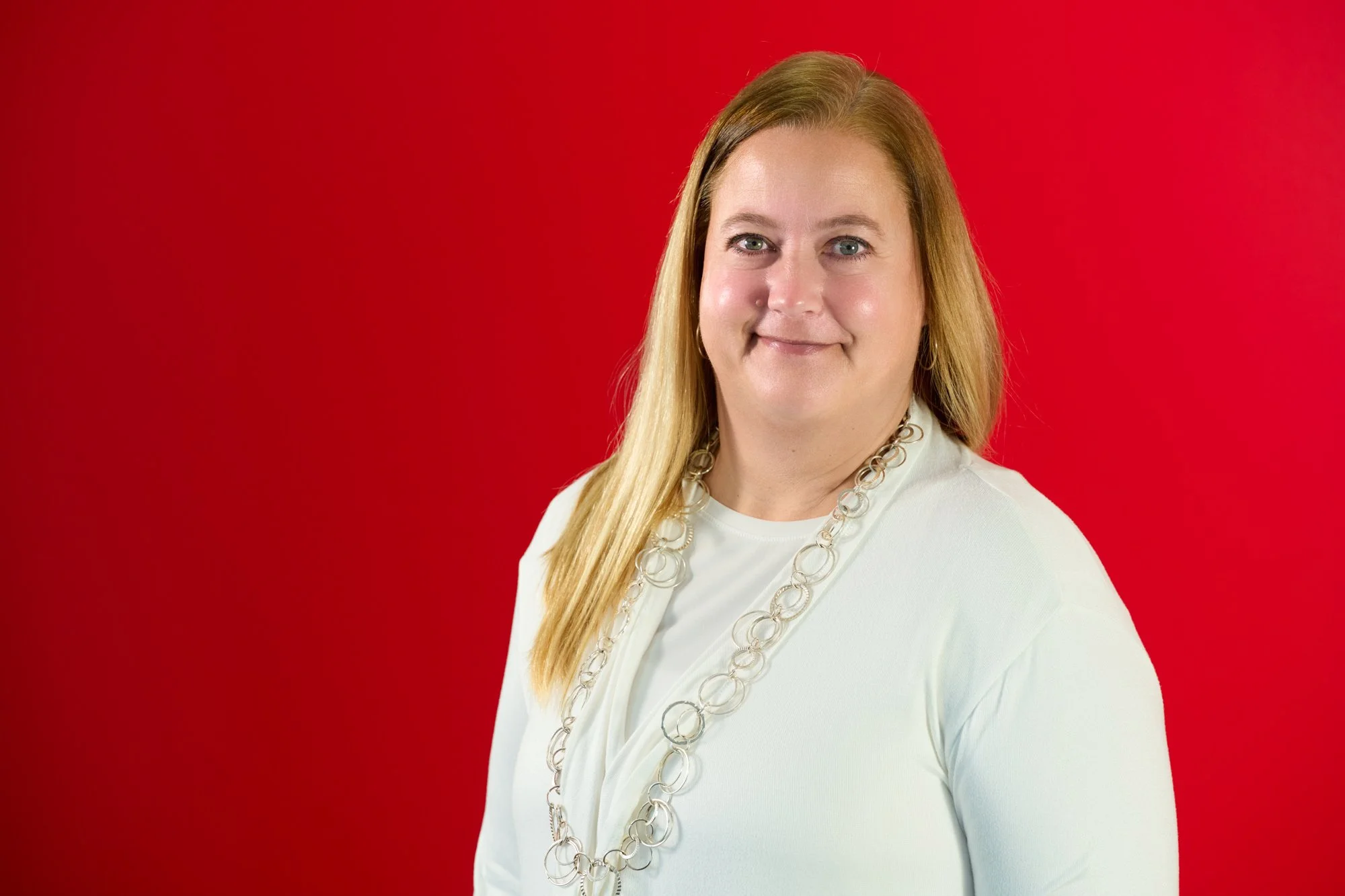 Portrait of a woman with long blonde hair wearing a white top and a long silver chain necklace, standing against a red background.