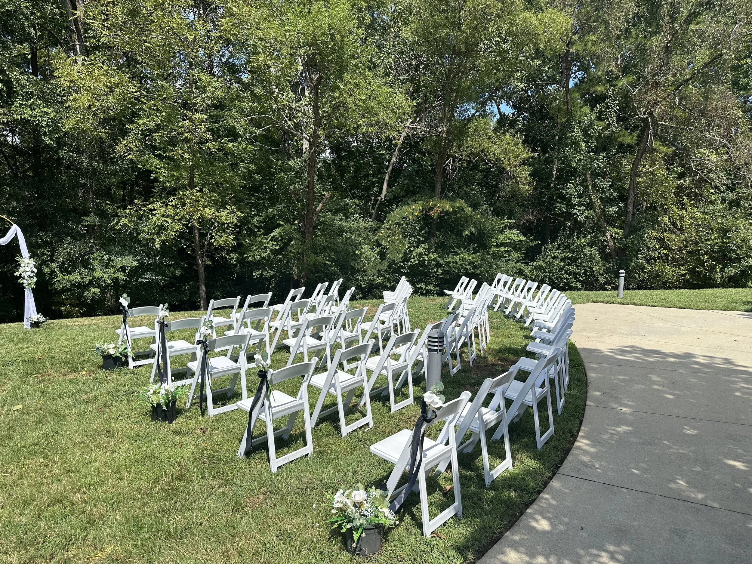Empty white folding chairs arranged in rows for a wedding ceremony outdoors on a grassy area next to a concrete sidewalk, with a wooded background and a white fabric arch decorated with flowers on the left.