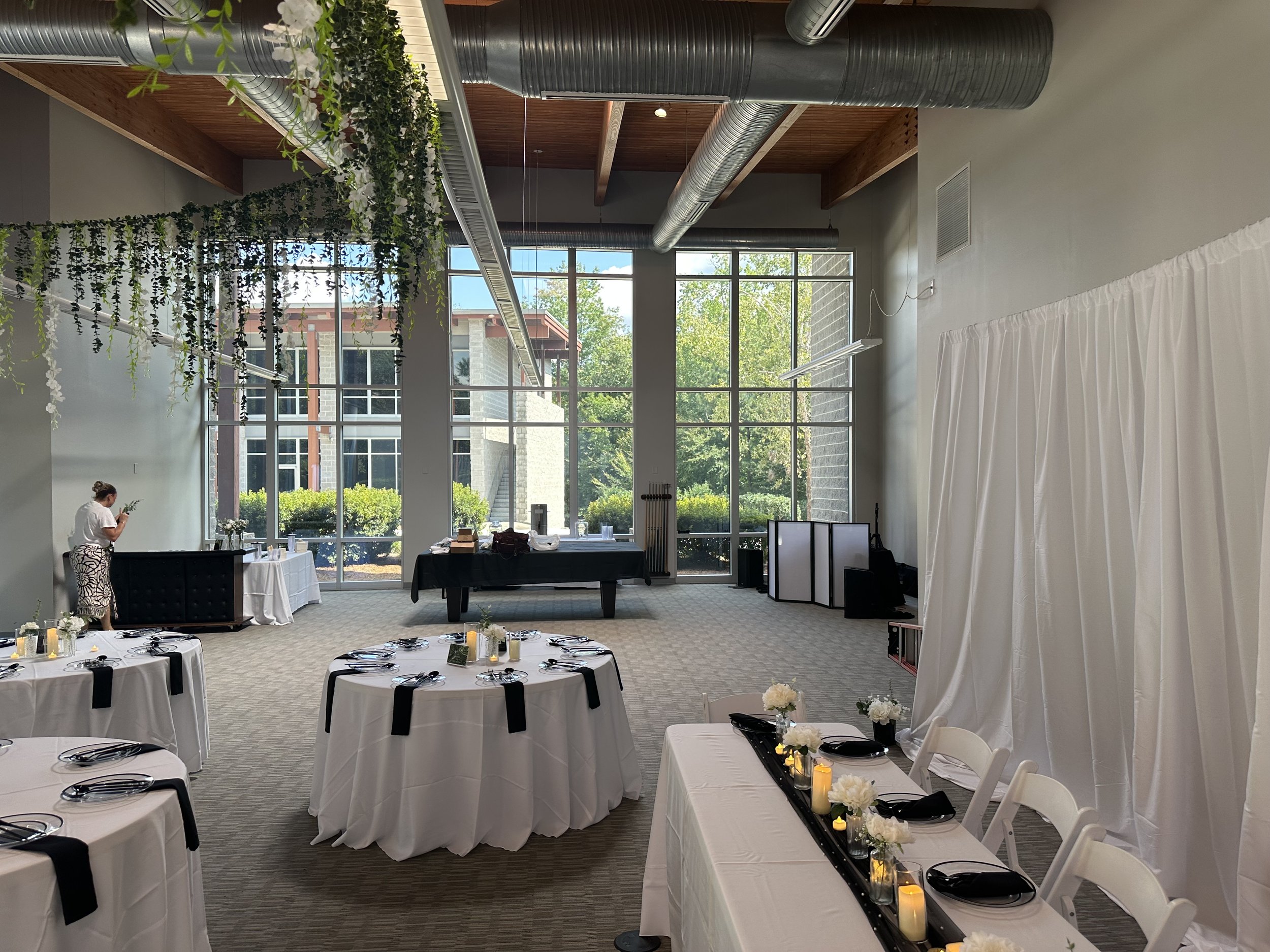 Indoor event space with round tables covered in white tablecloths and black napkins, decorated with candles and white flowers. Large windows reveal trees and a building outside. Hanging greenery and white flowers decorate the ceiling, which has expos