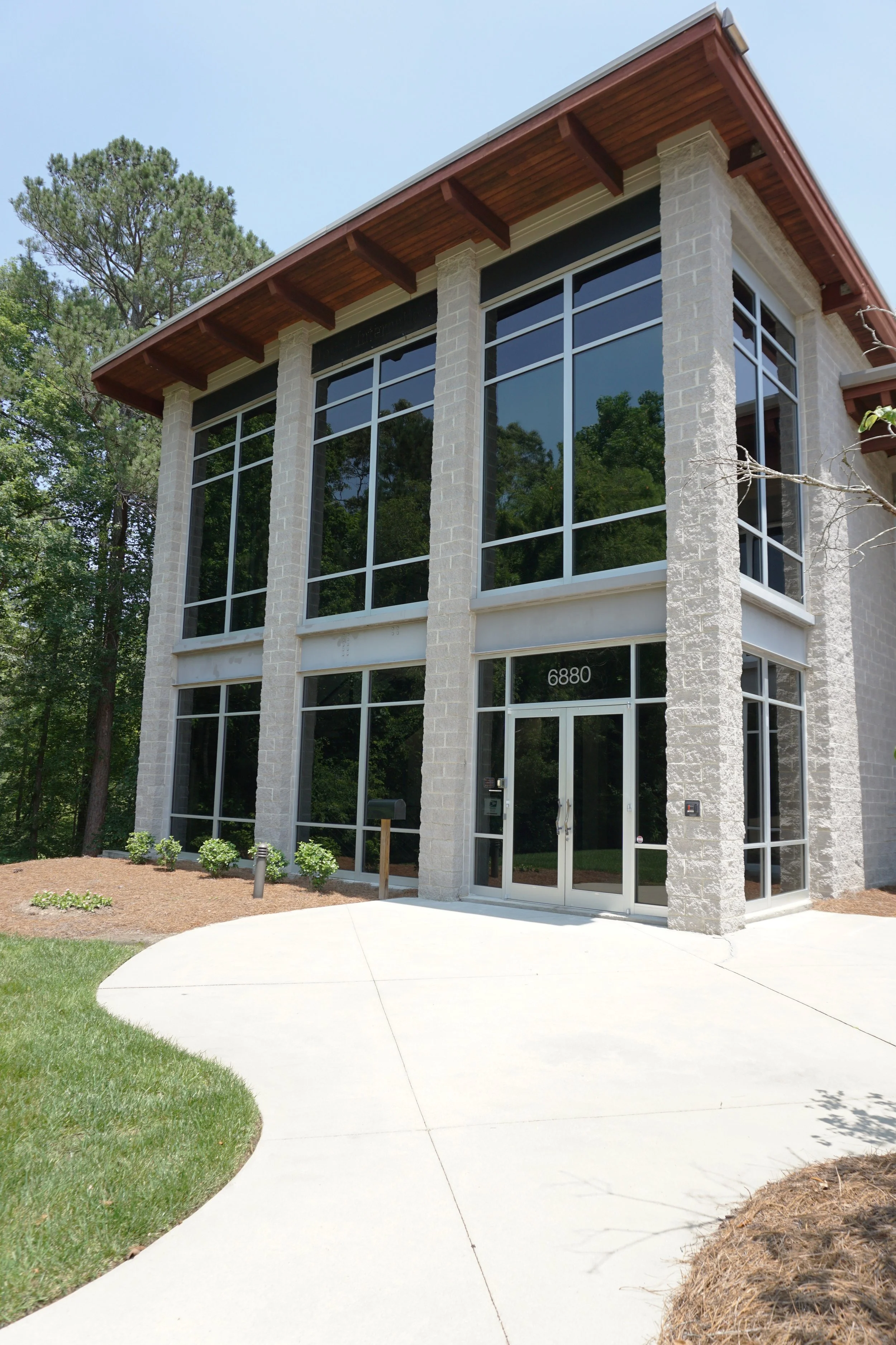Modern two-story office building with large glass windows, brick columns, and a wooden roof overhang. The building number is 6880, and there is a sidewalk leading to the entrance, surrounded by landscaped grass and small bushes.