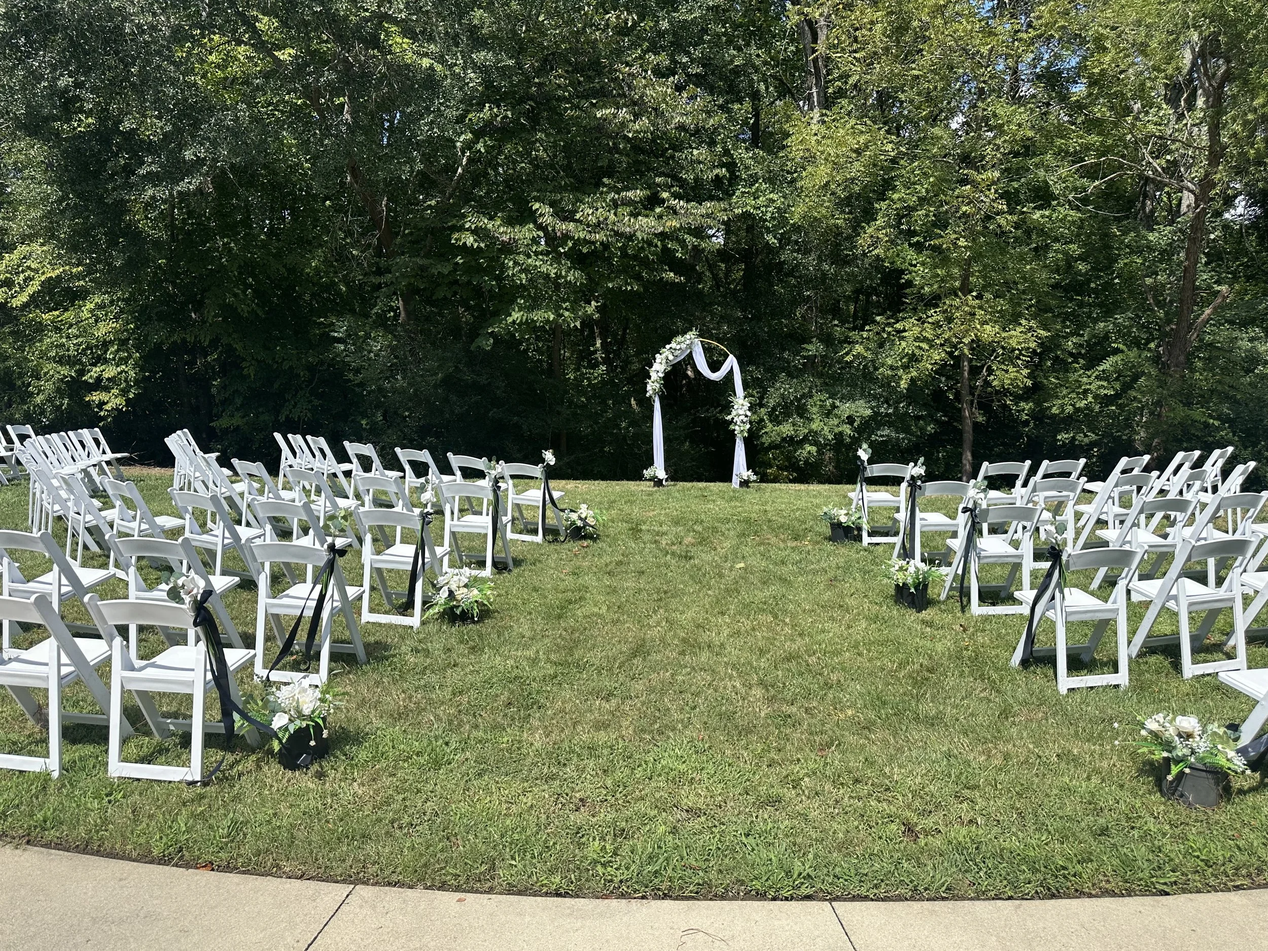Outdoor wedding ceremony setup with white folding chairs arranged on grass, decorated with flowers and ribbons, facing an arch decorated with white flowers and fabric, surrounded by green trees.