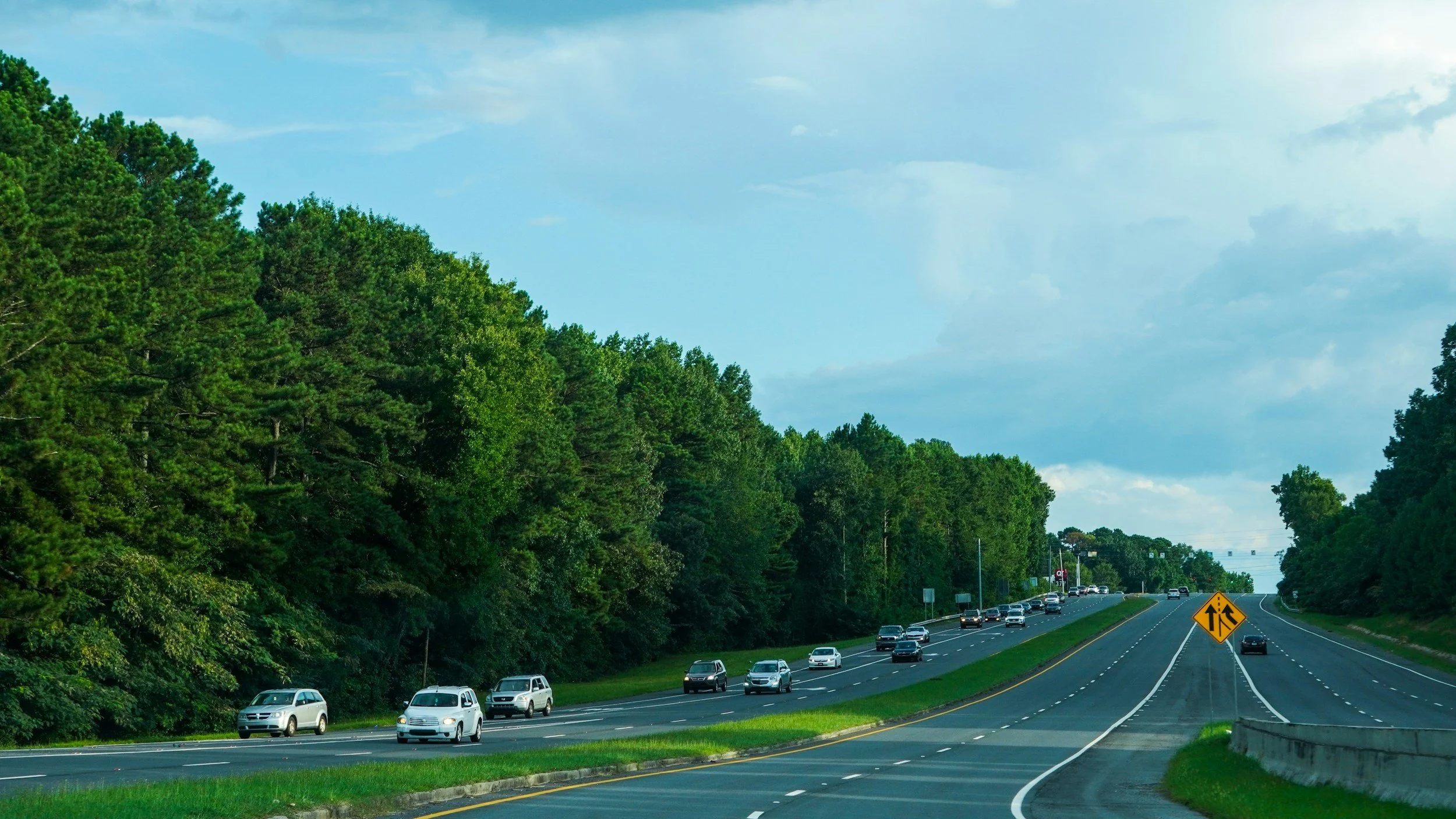 A multi-lane highway with cars traveling in both directions, bordered by lush green trees on the left and right, with a sky filled with clouds overhead, and a yellow roadwork sign on the right.