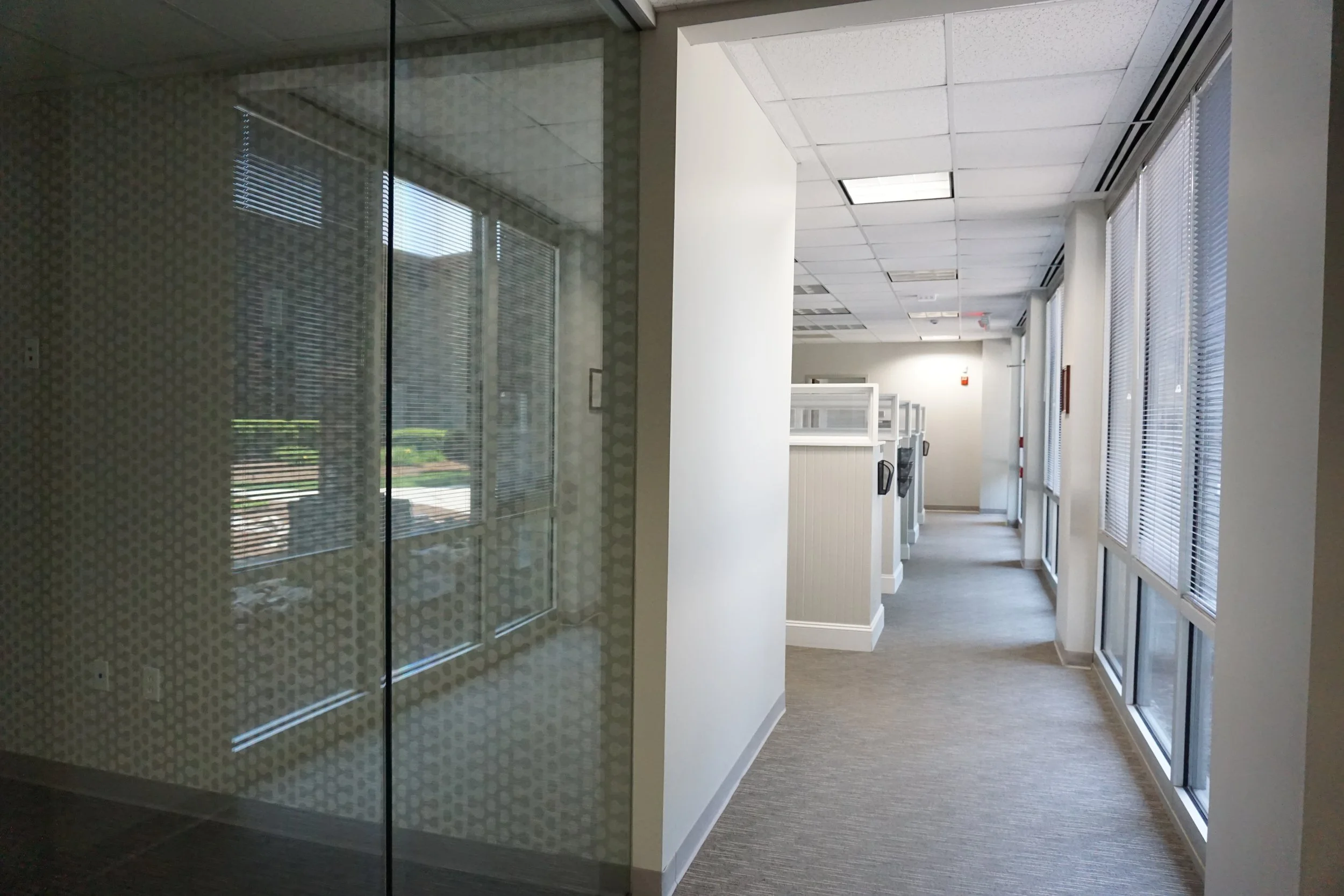 Empty office hallway with cubicles, windows with blinds on the right, and a glass door on the left, illuminated by fluorescent ceiling lights.
