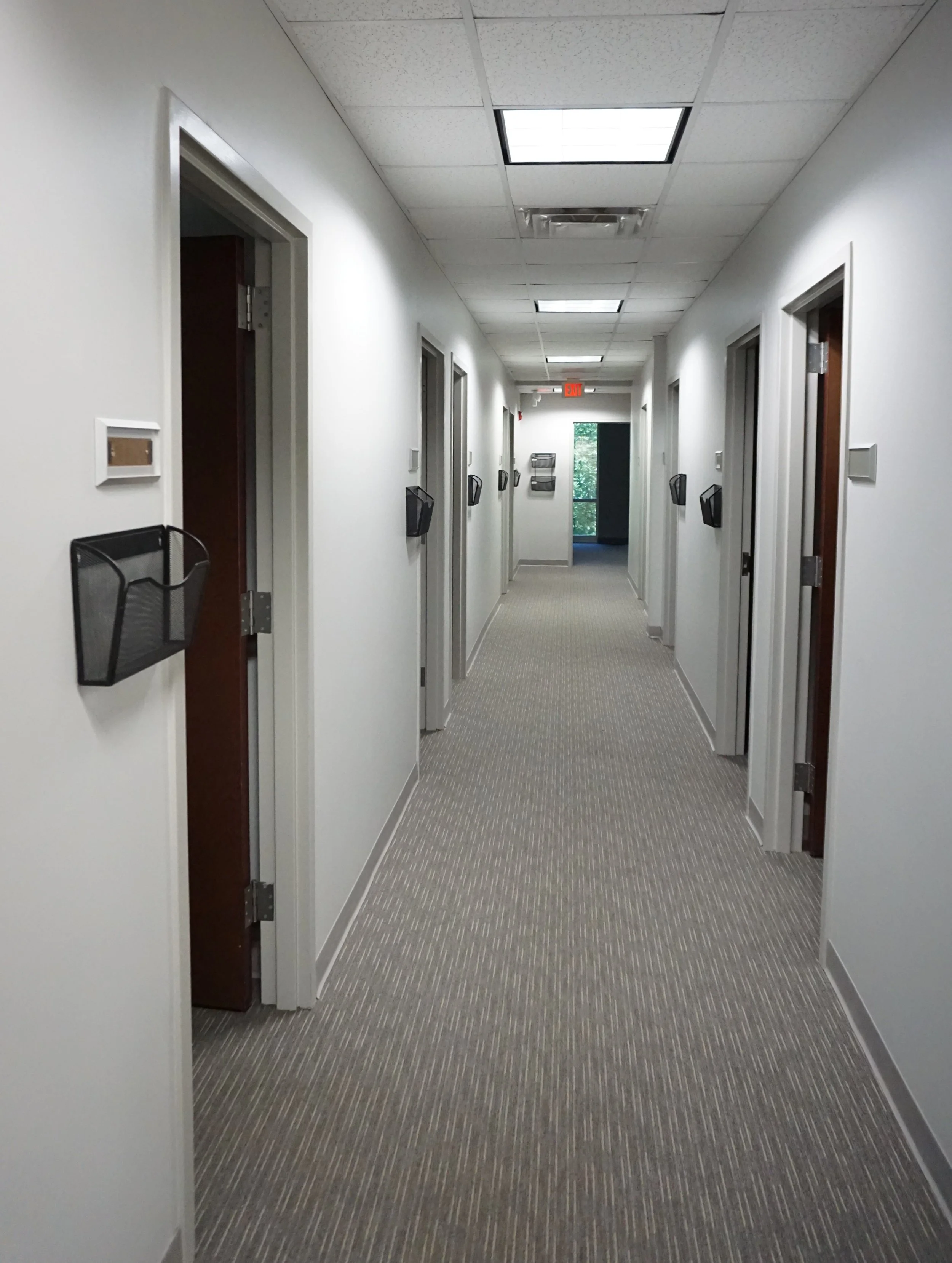Empty office hallway with white walls, beige carpet, and closed doors with black wall-mounted file holders.