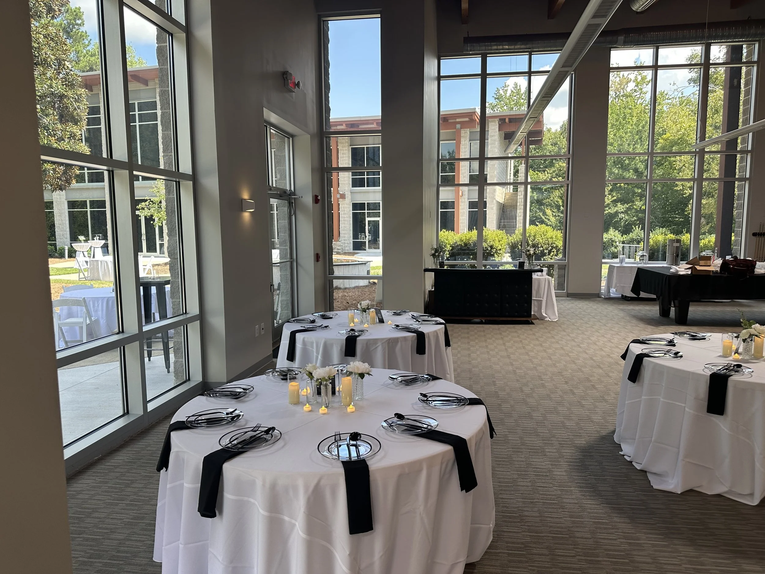 Interior of a modern event space with large windows, round tables with white tablecloths, black napkins, and place settings, and a view of outdoor tables and greenery.