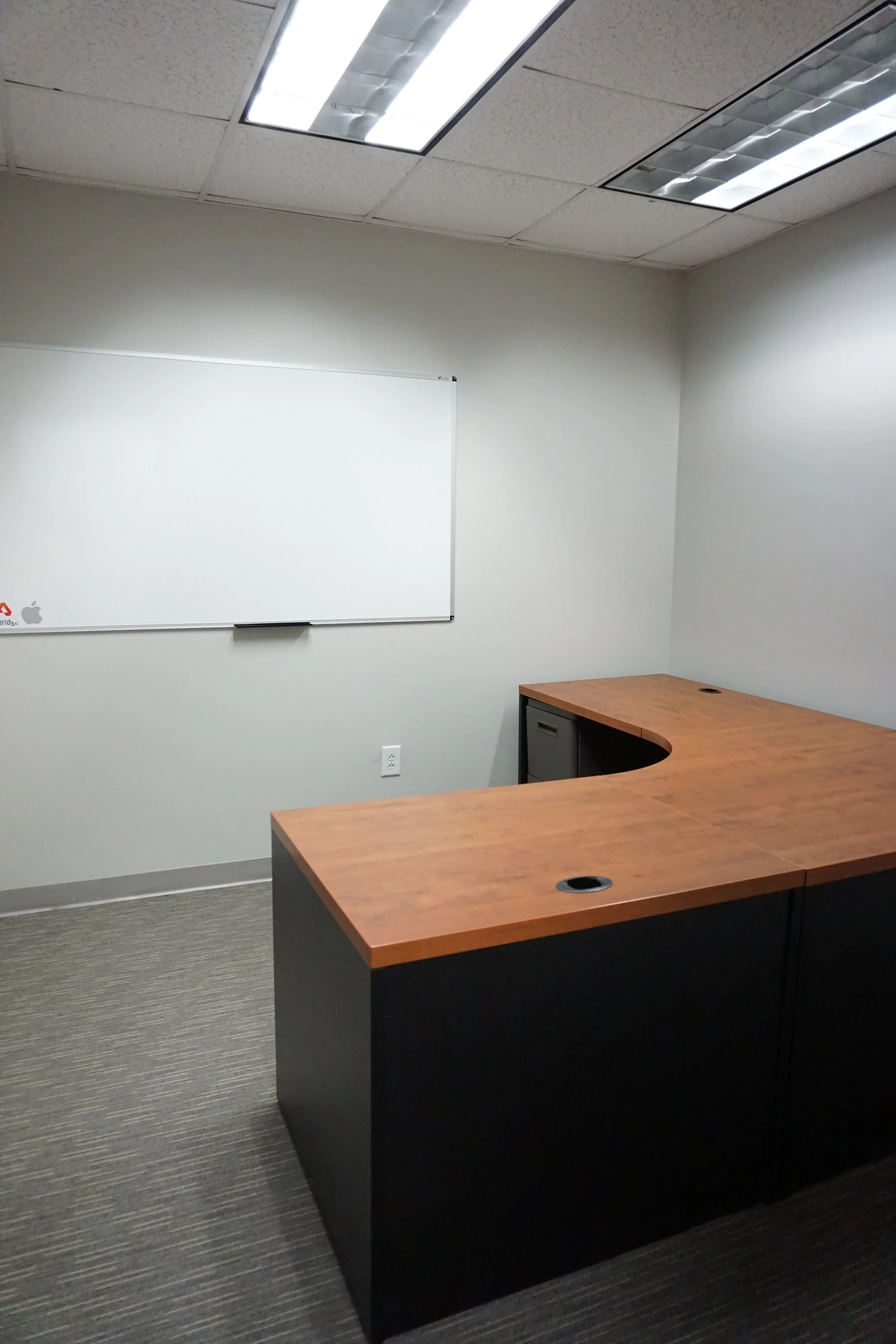 Empty office room with a wooden L-shaped desk, a whiteboard on the wall, and ceiling fluorescent lights.