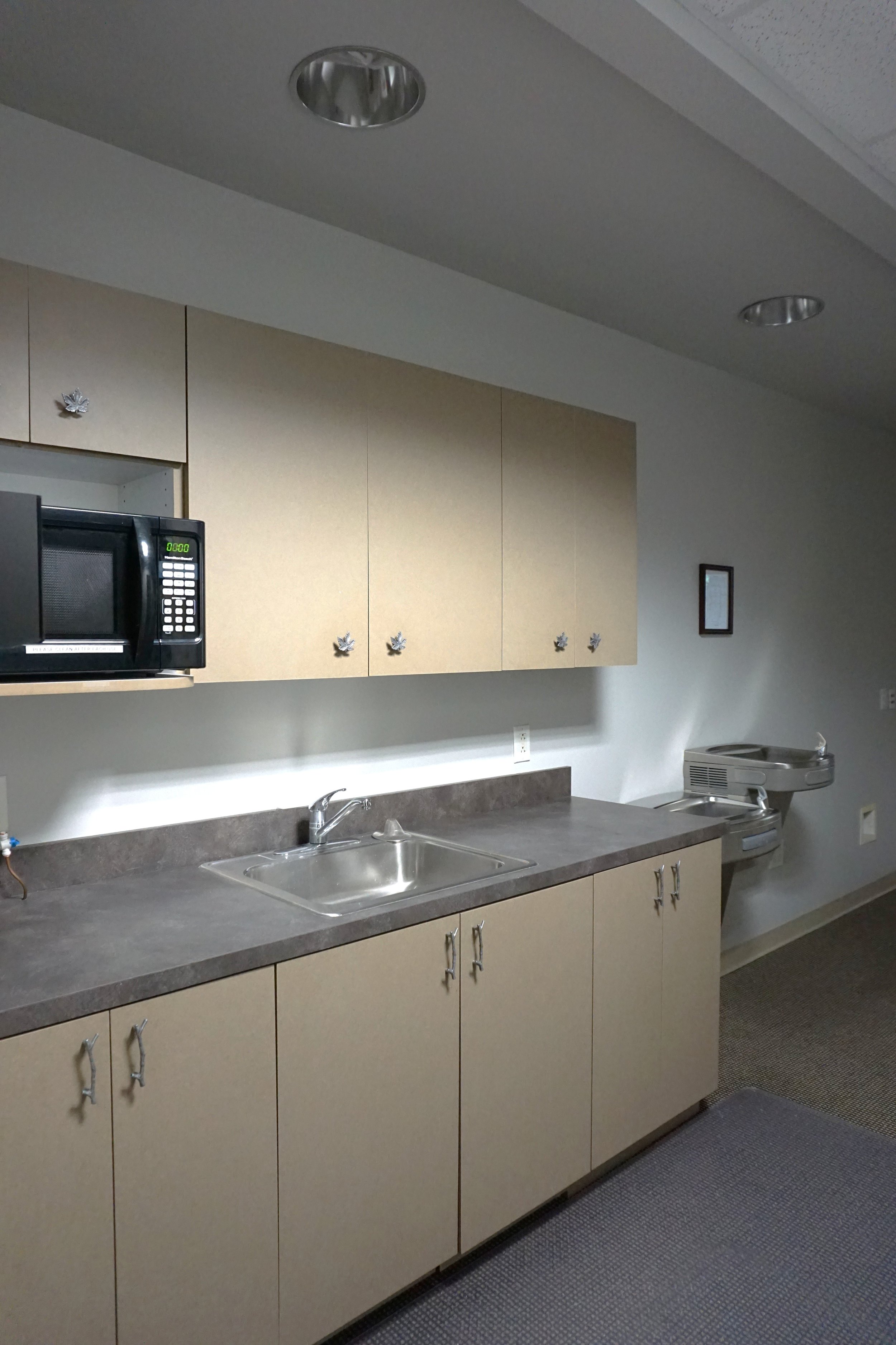 Empty kitchenette with beige cabinets, gray countertop, stainless steel sink, microwave, and a drinking fountain in the background.