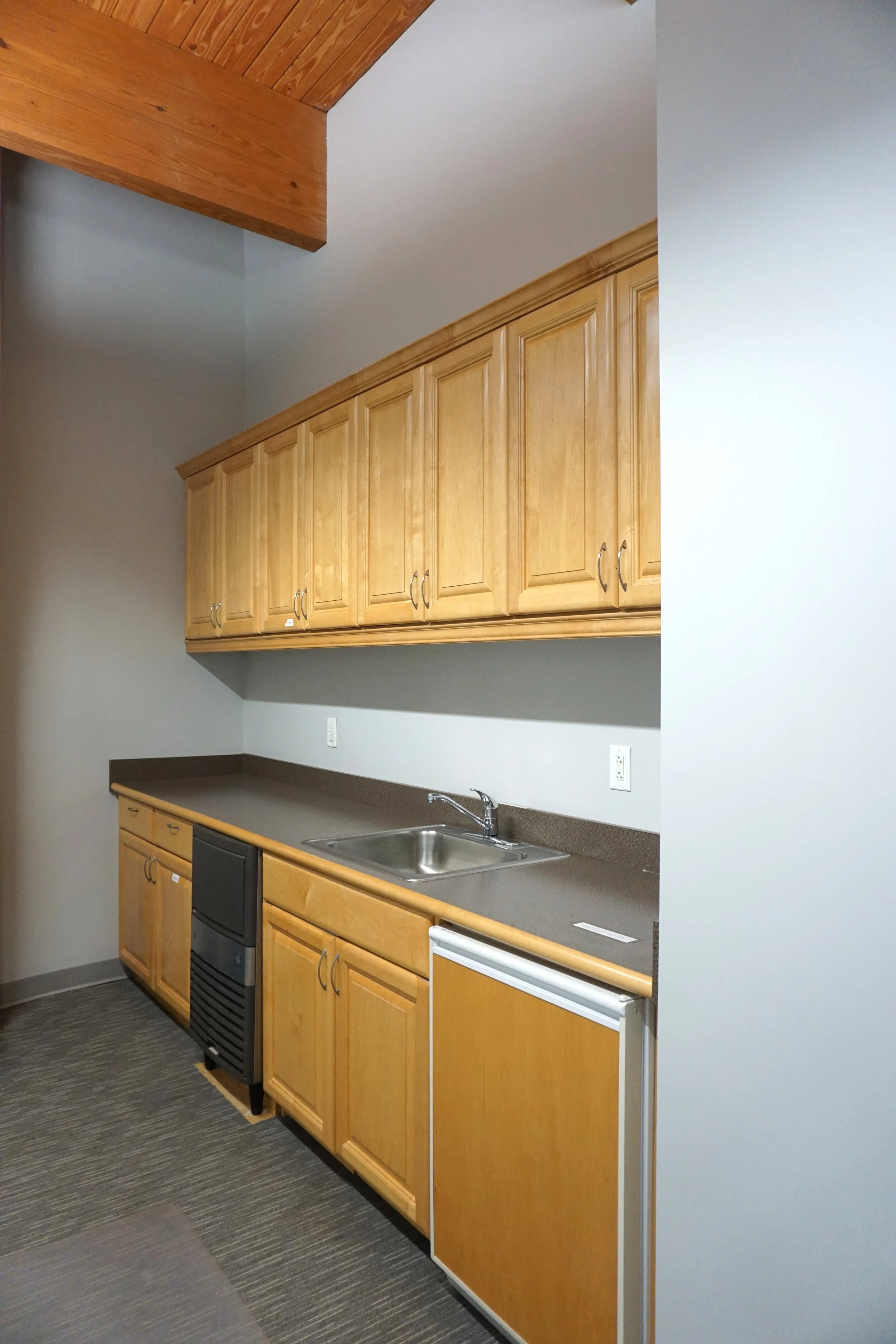 Empty kitchenette with light wood cabinets, a small refrigerator, a sink, and a countertop in an office or break room area.