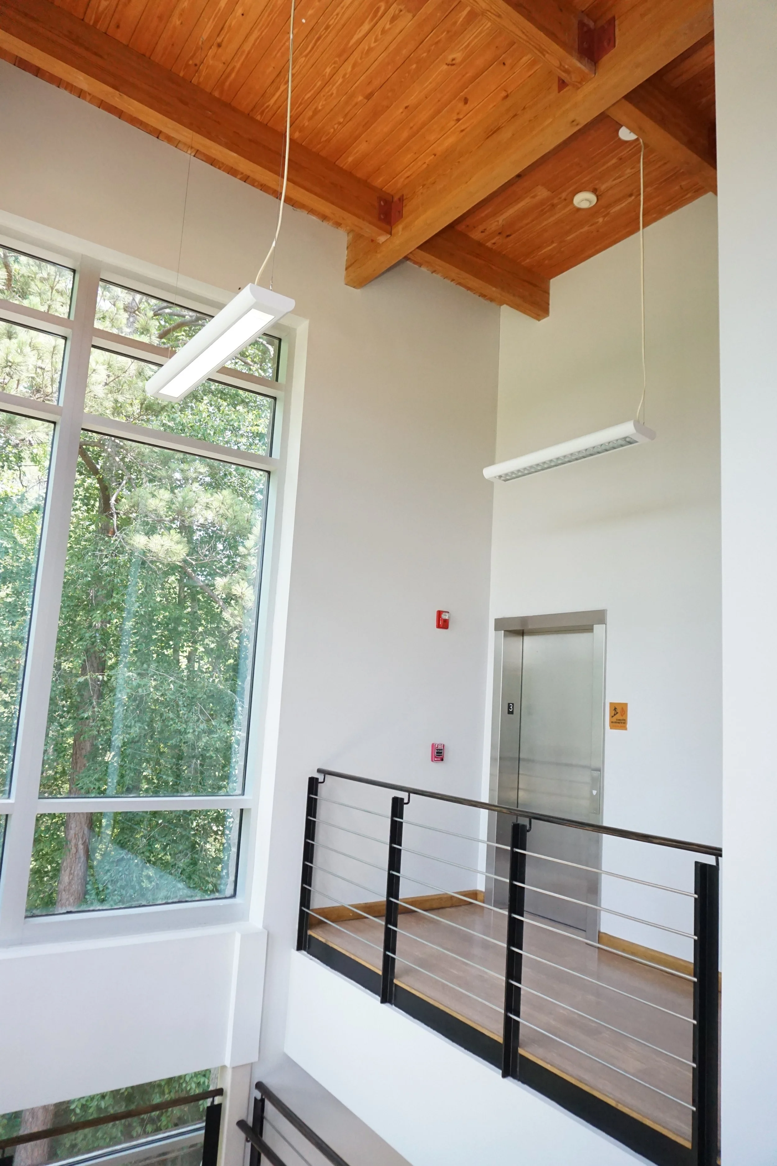 Interior of a modern building featuring a wooden ceiling with exposed beams, large window with views of green trees, and an elevator door. Two white hanging light fixtures are suspended from the ceiling, and a black metal railing surrounds a small la