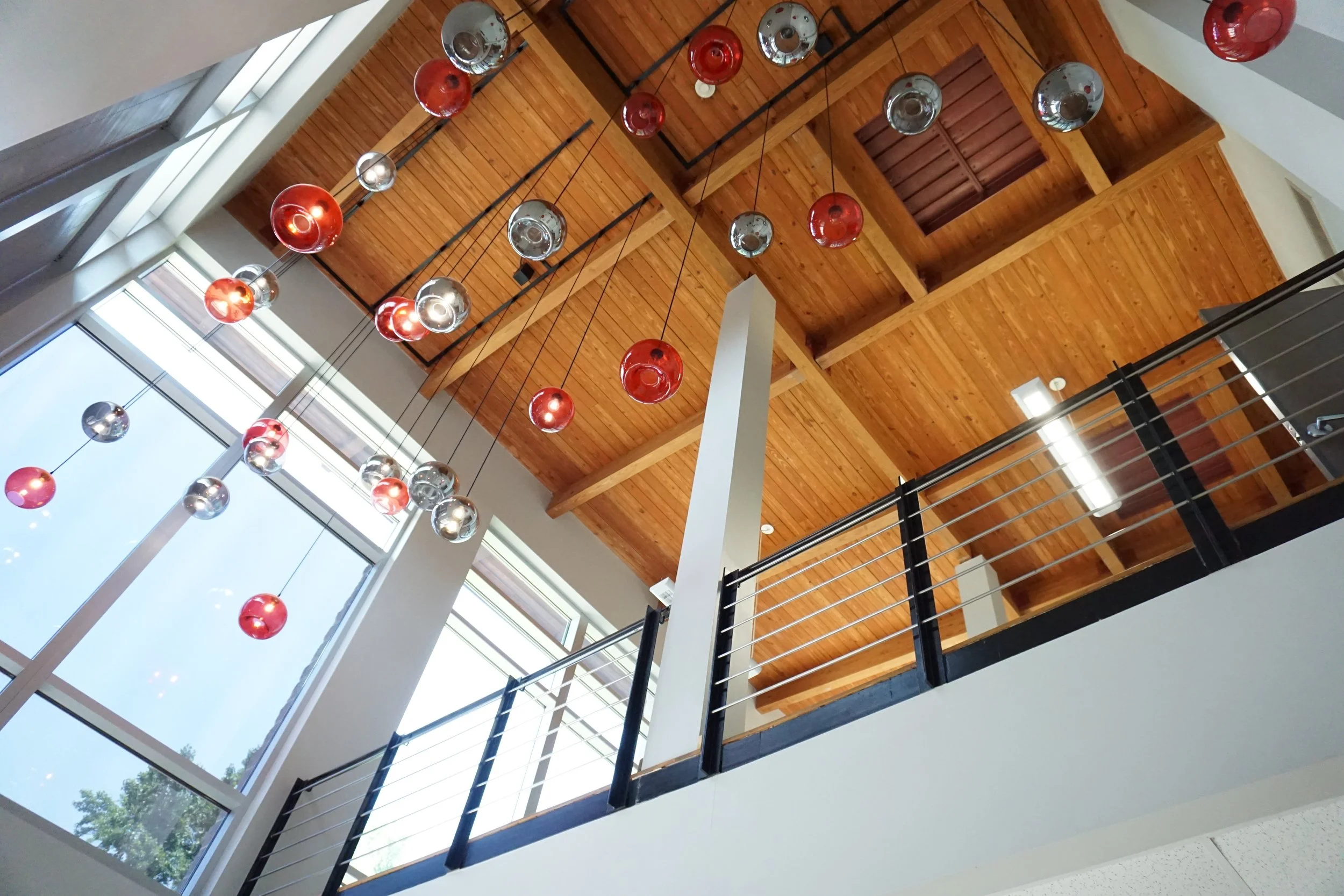 Interior view of a modern building with a wooden ceiling, hanging spherical red and silver light fixtures, large windows, and a black metal railing.