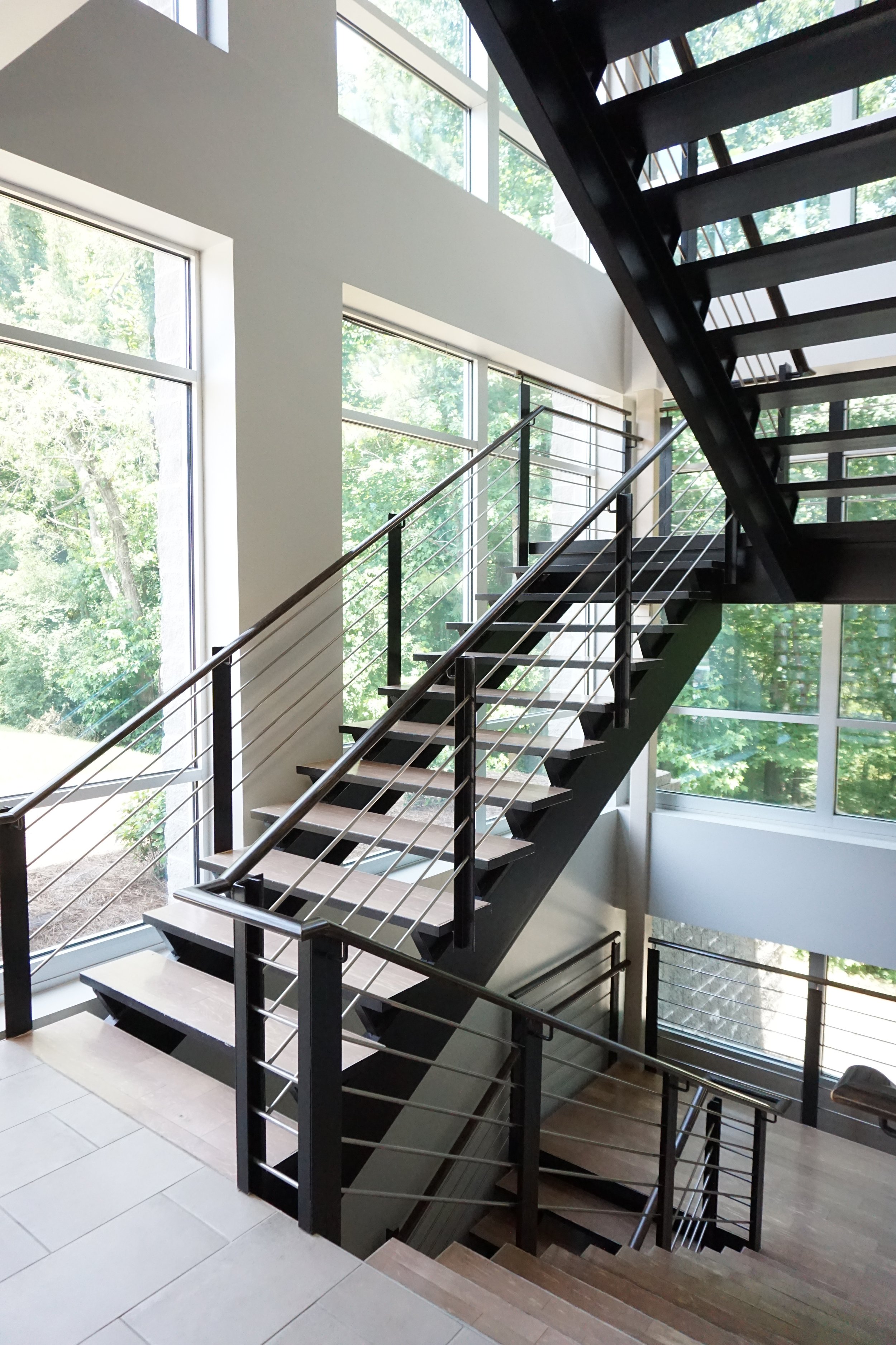 Interior of a modern building featuring a staircase with black steps and metal railings, large floor-to-ceiling windows letting in natural light, and a view of green trees outside.