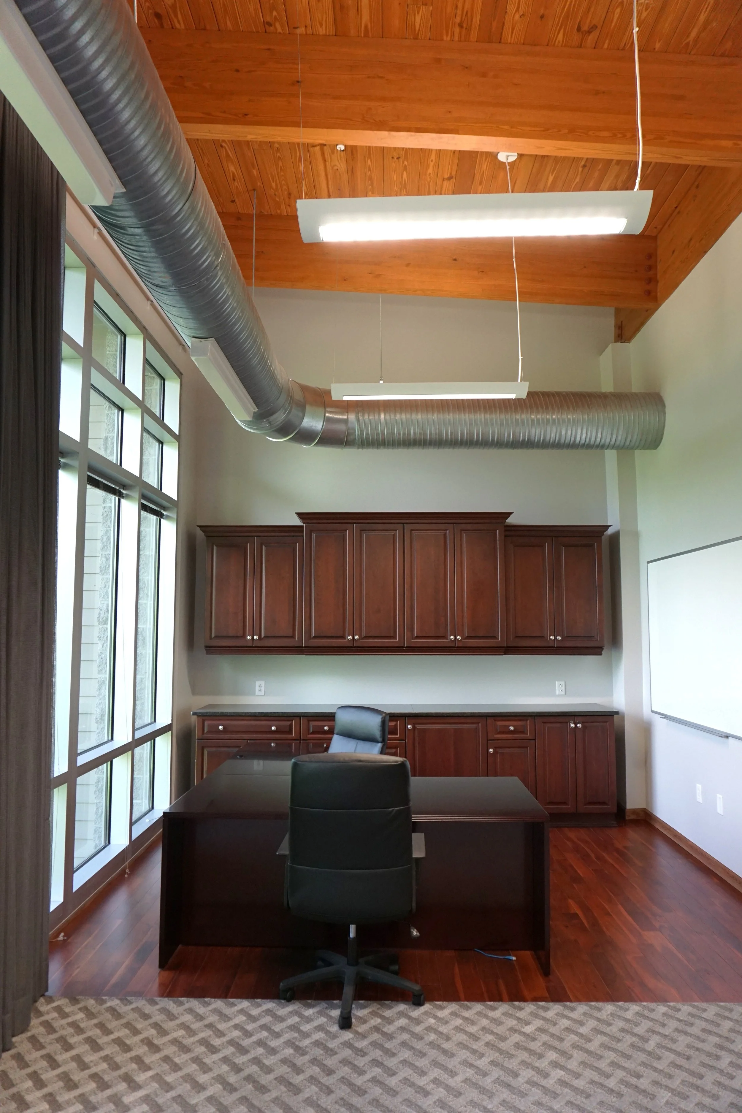 A modern office with dark wooden desk and cabinets, black office chair, large windows with curtains, wood-paneled ceiling, exposed ventilation ducts, and a whiteboard on the wall.