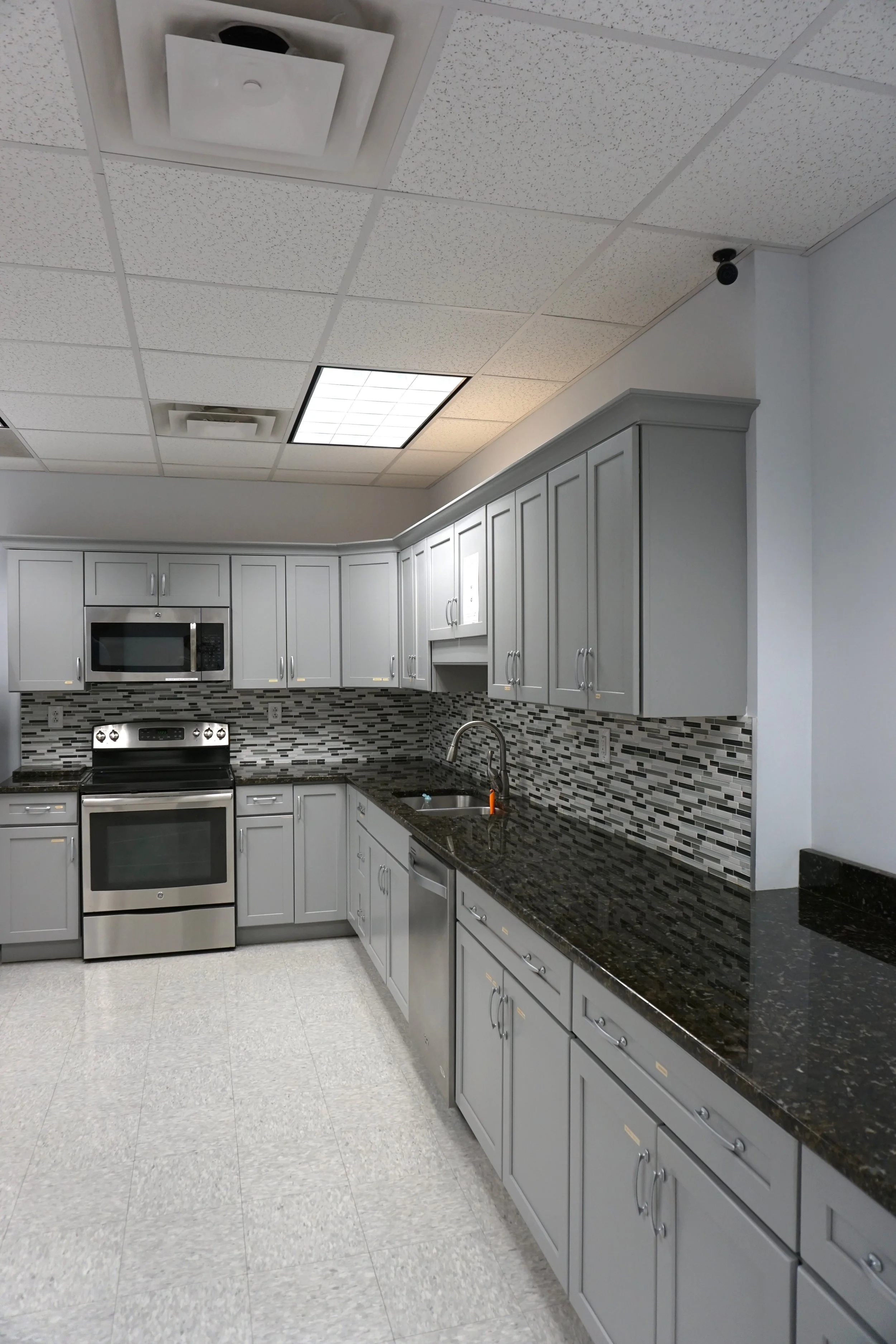 A modern kitchen with gray cabinets, black granite countertops, a mosaic tile backsplash, stainless steel appliances, including an oven, microwave, and dishwasher, and a ceiling with fluorescent lights.