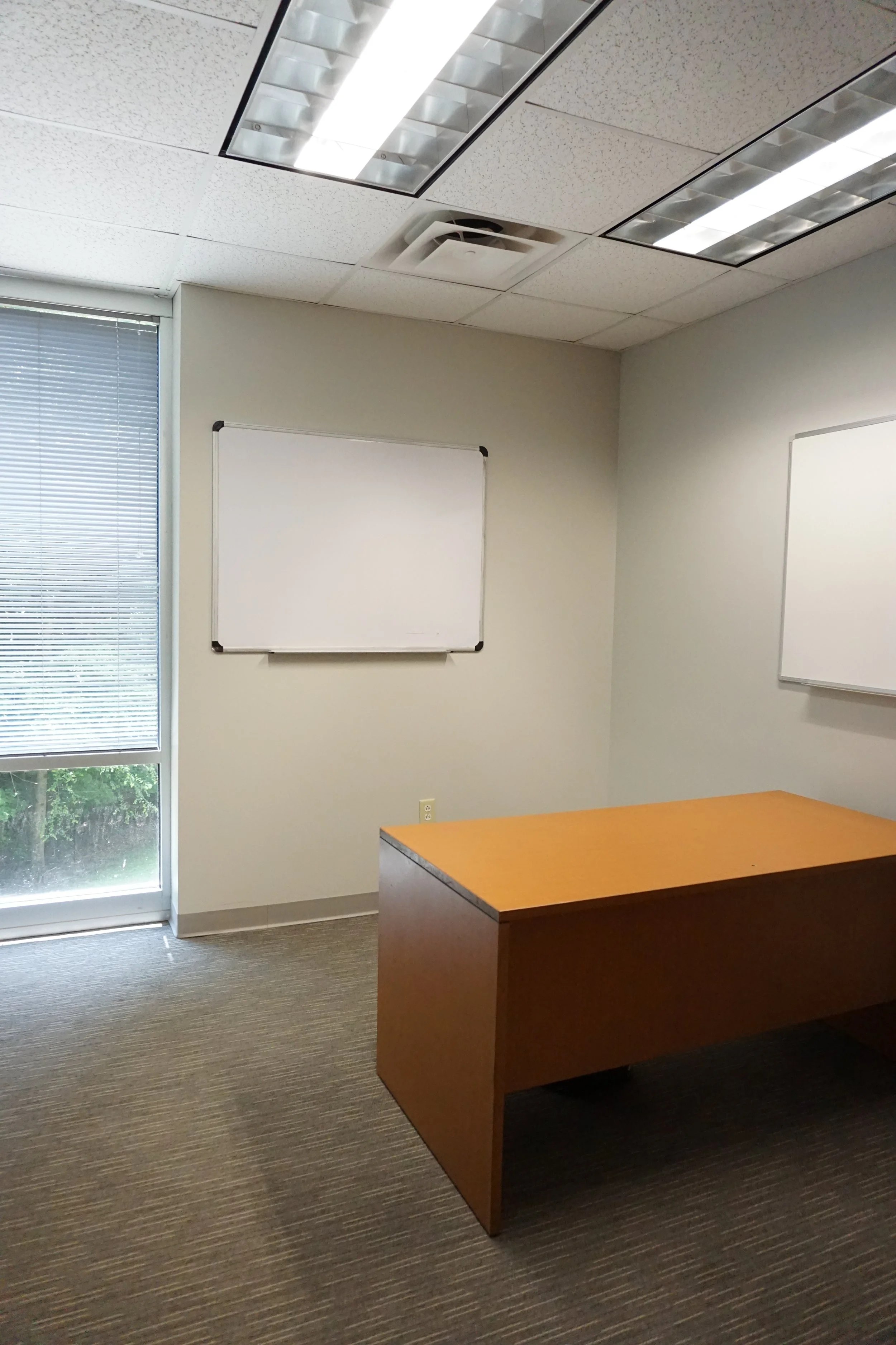 Empty office room with a wooden desk, two whiteboards on the walls, a window with blinds, and a ceiling light panel.