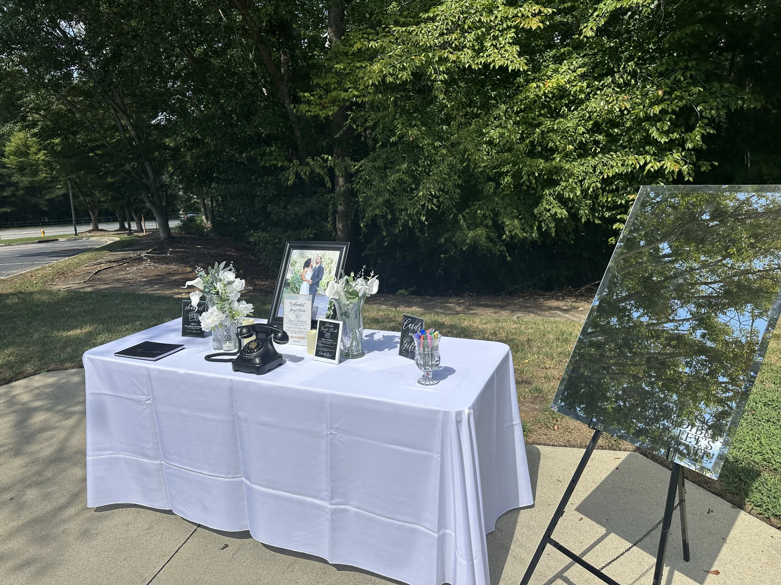 Outdoor memorial table with framed photograph of a couple, flowers, and signs, set against a backdrop of trees and a sidewalk.