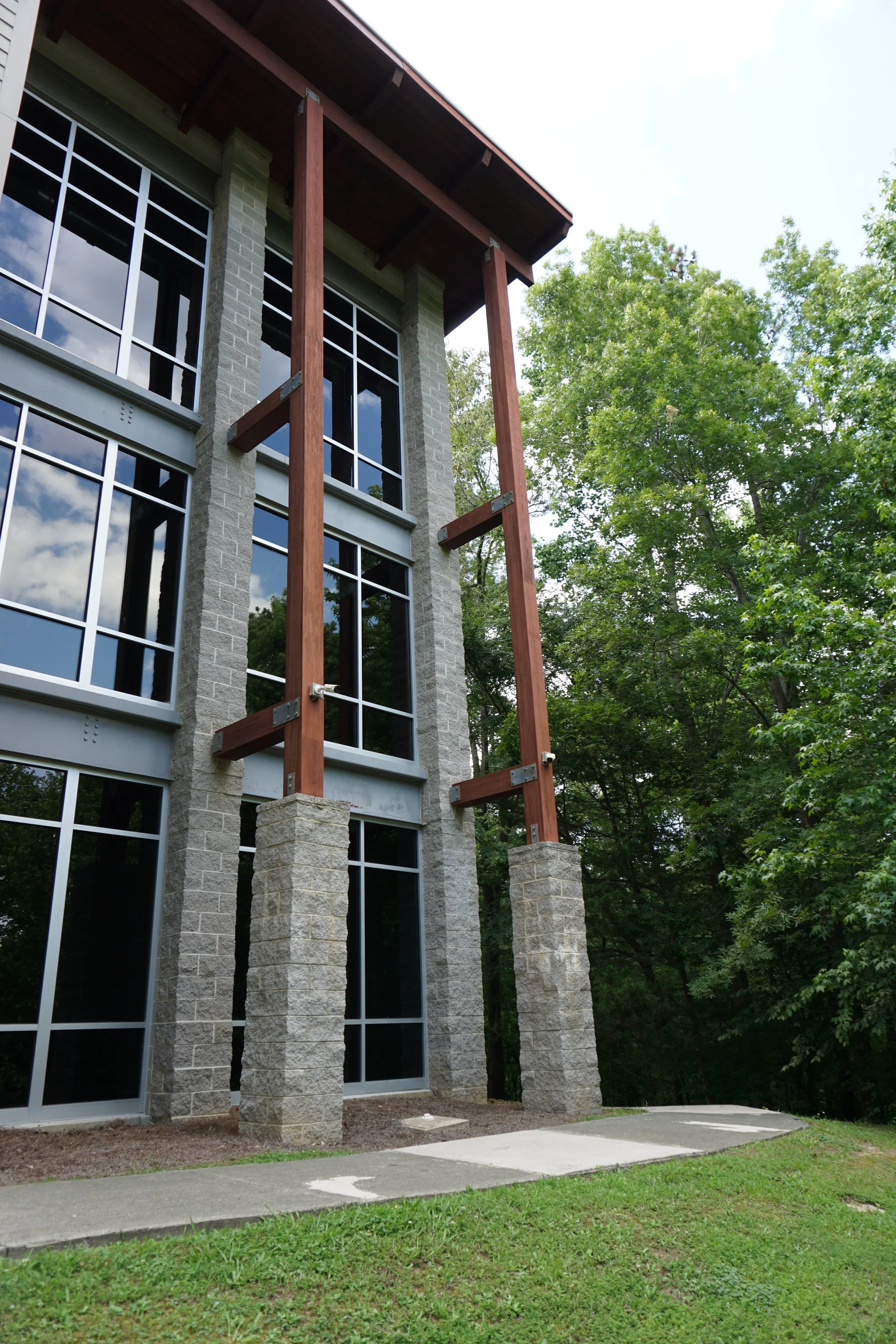 The image depicts the exterior of a modern building under construction, showing large glass windows, gray brick walls, and exposed wooden beams supported by stone pillars.