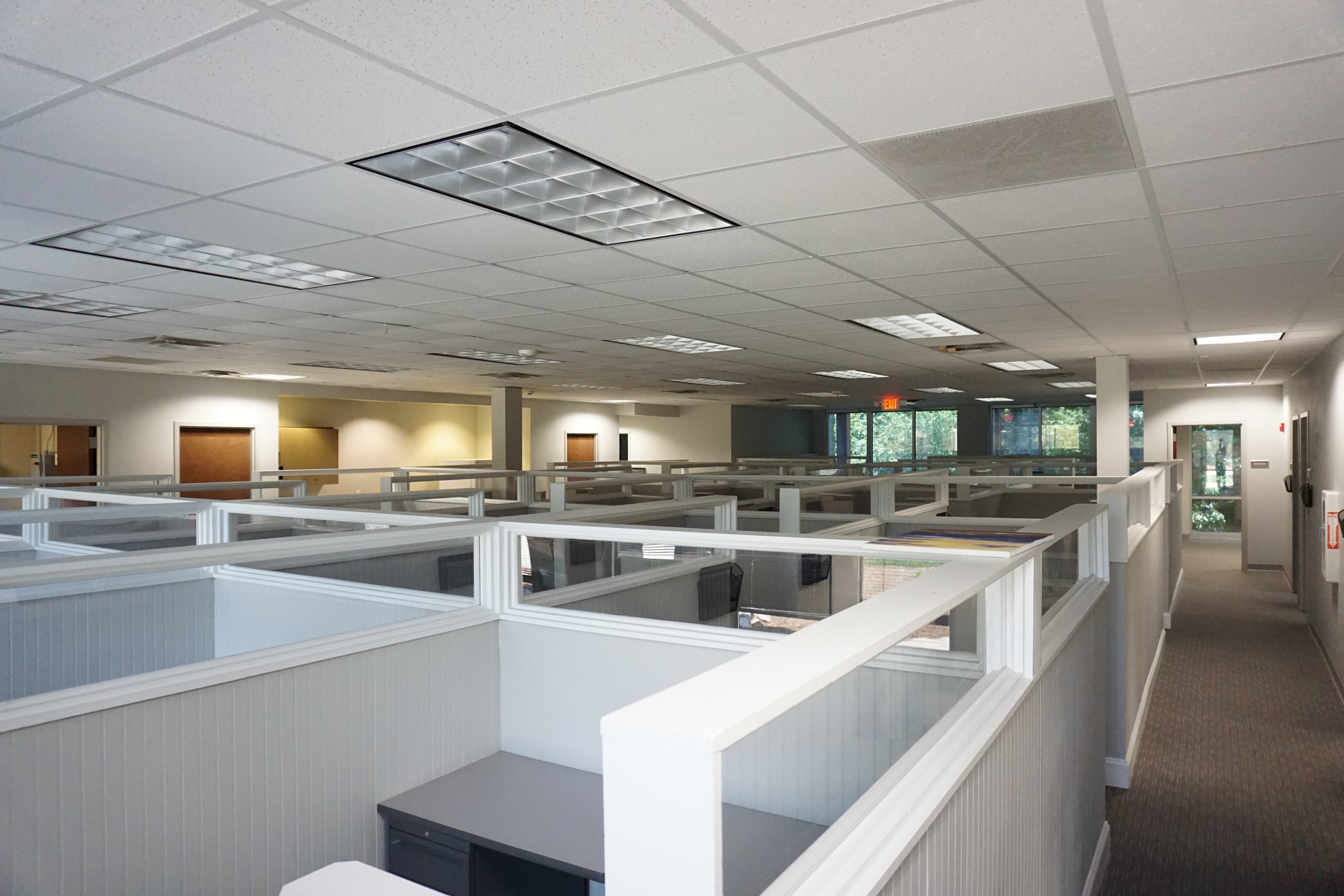 Empty office space with cubicles, fluorescent ceiling lights, and glass windows in the background.