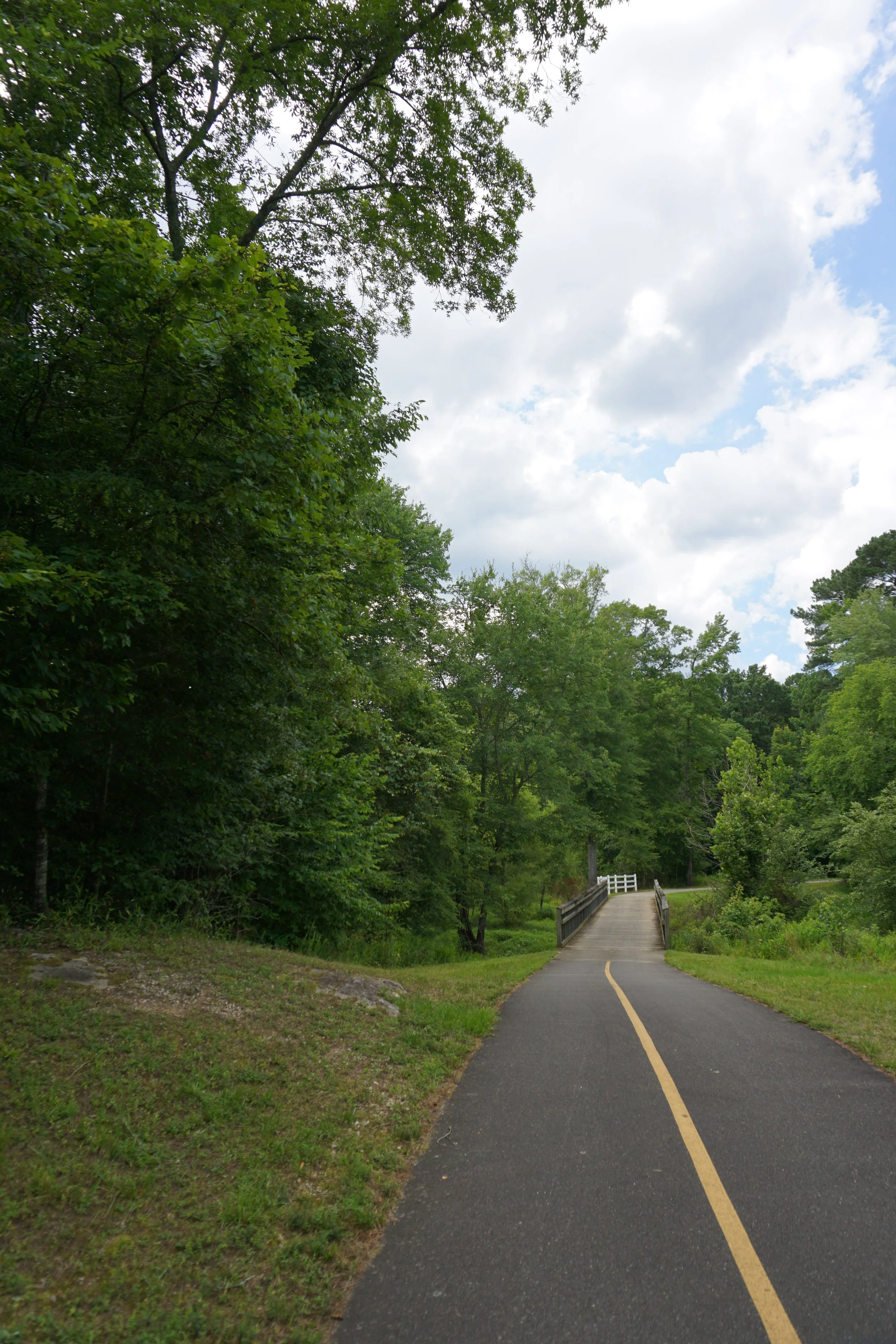 A paved walking trail with a yellow center line, leading to a small wooden bridge surrounded by green trees and a partly cloudy sky.