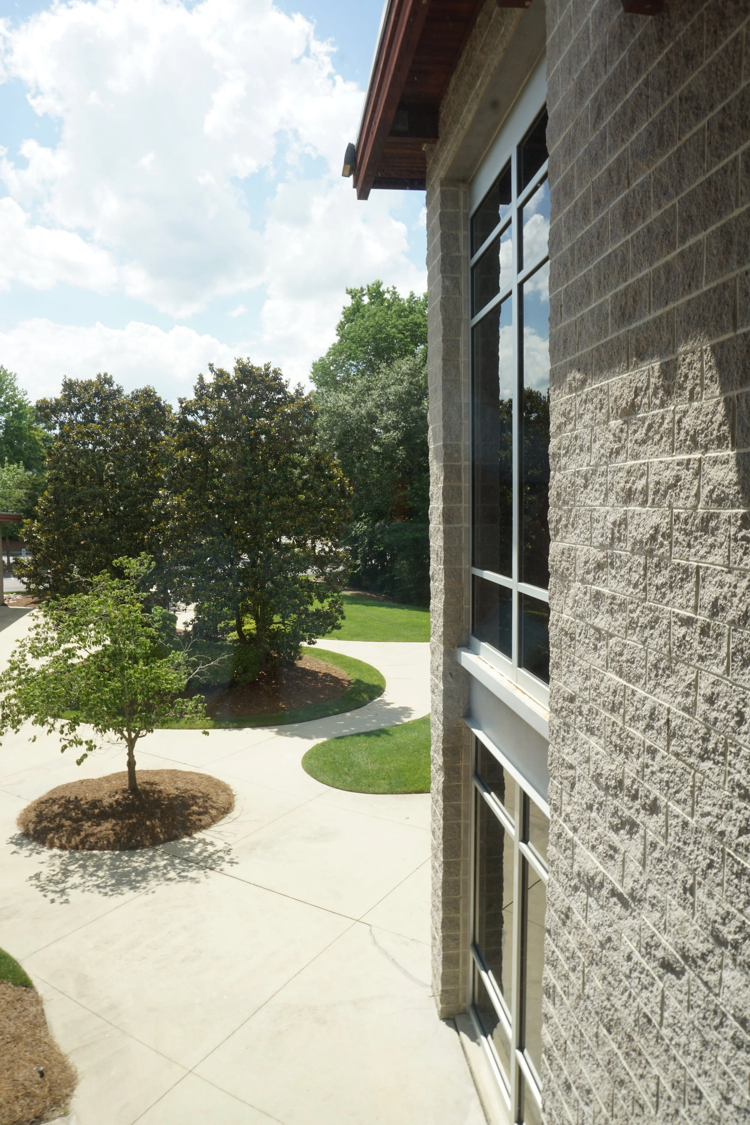 View of a modern building with large glass windows on a brick wall, with landscaped trees and a curved sidewalk on a sunny day with blue sky and some clouds.
