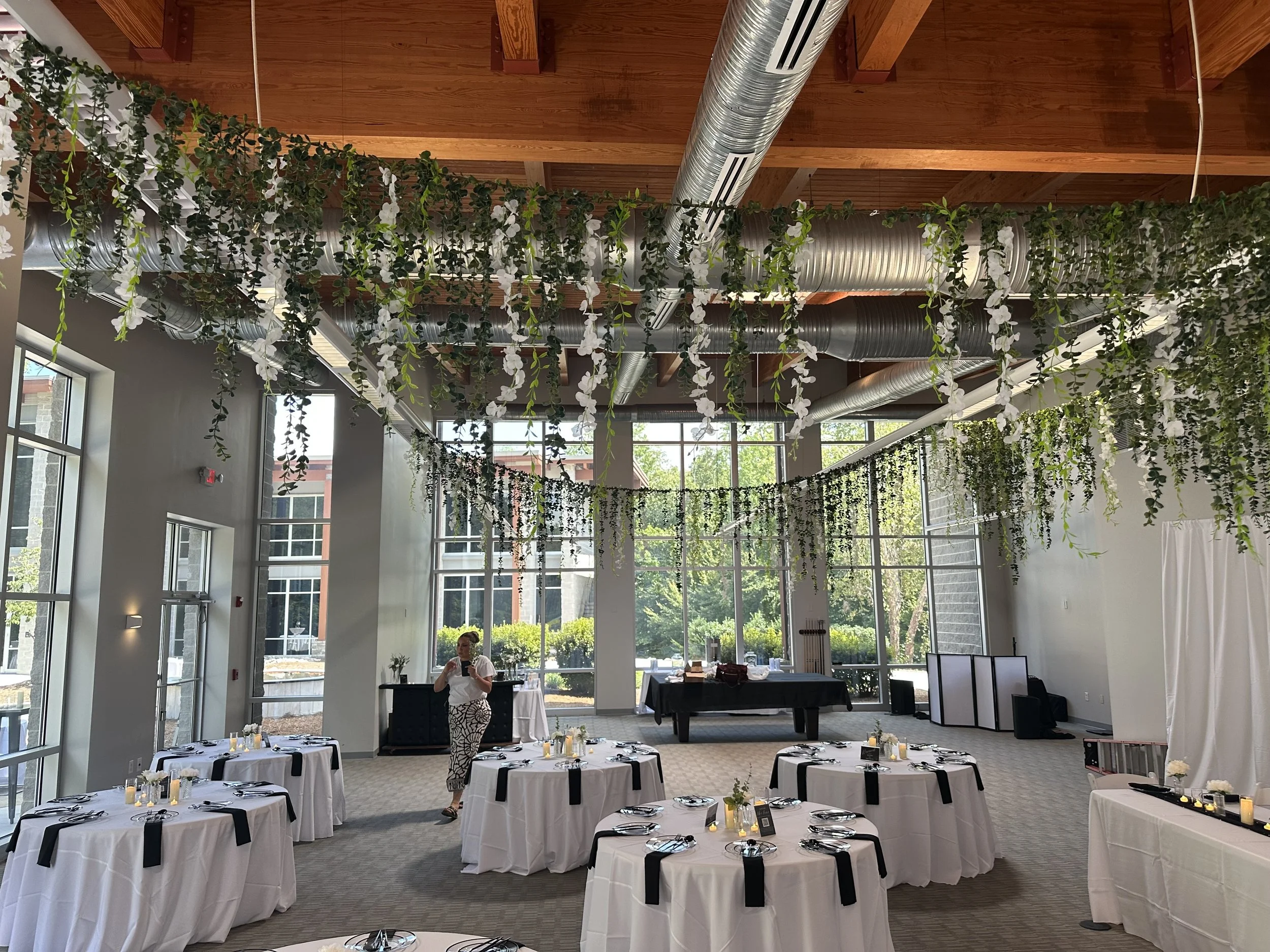 Event space with high ceilings, large windows, and hanging greenery and white flowers decor, tables set with black and white decorations, and a person standing near a table.