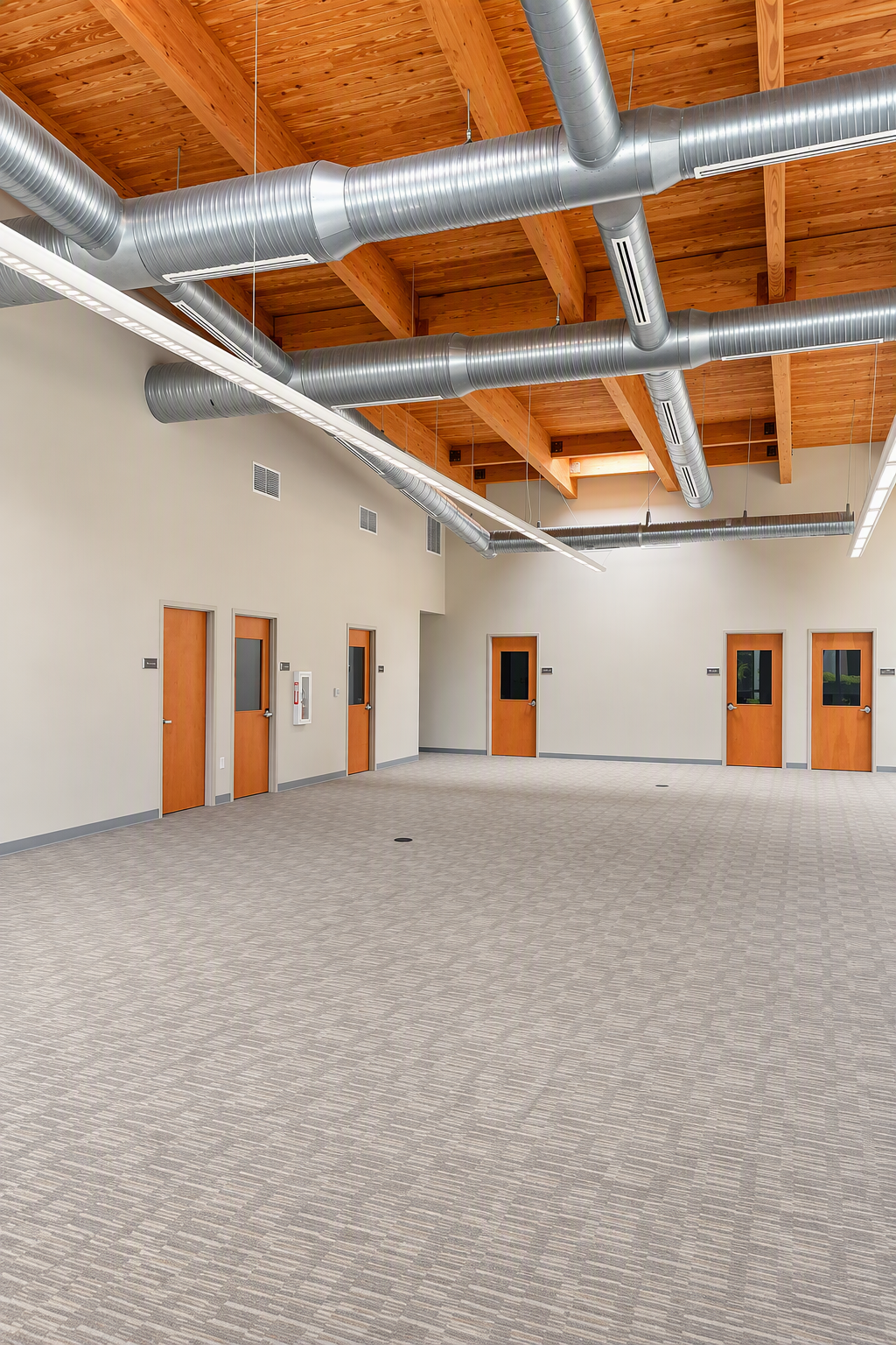Empty room with multiple wooden doors, beige walls, carpeted floor, exposed wooden ceiling beams, and visible silver air ducts.