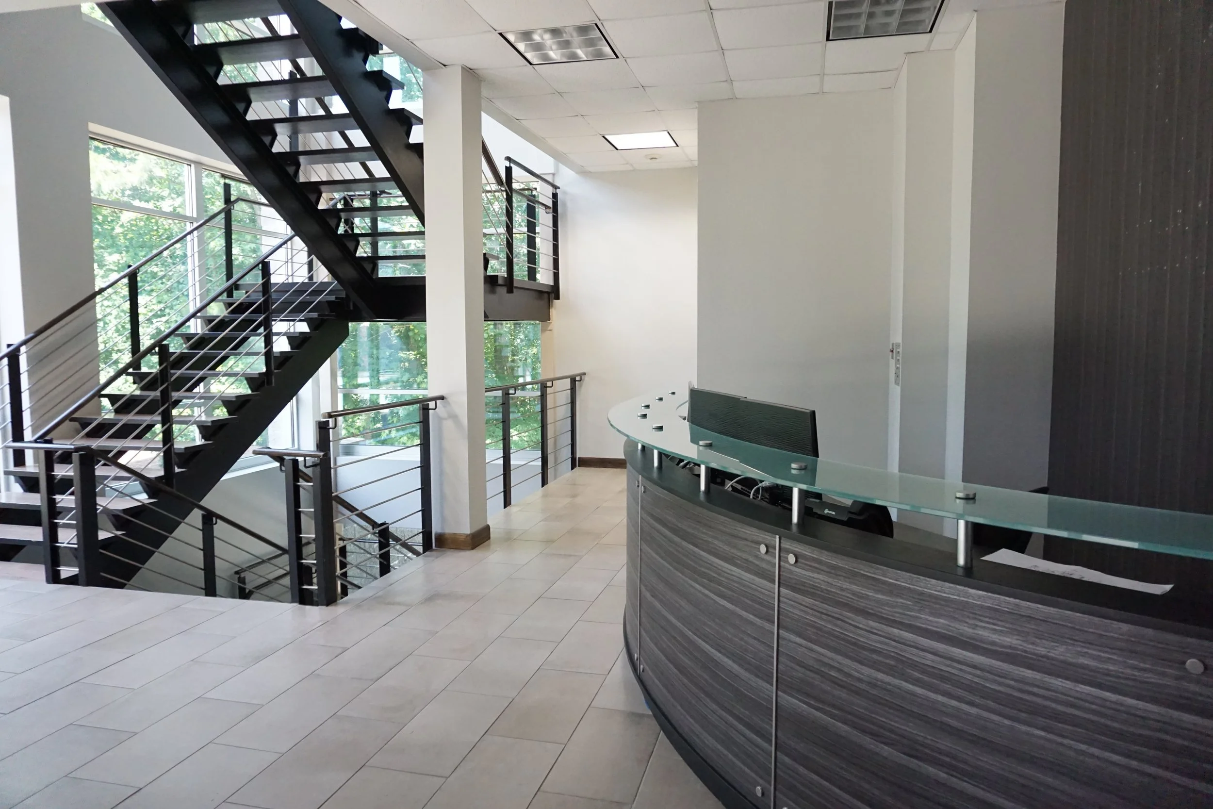Interior of a modern office with a curved reception desk, large windows, and a black metal staircase leading to an upper floor.