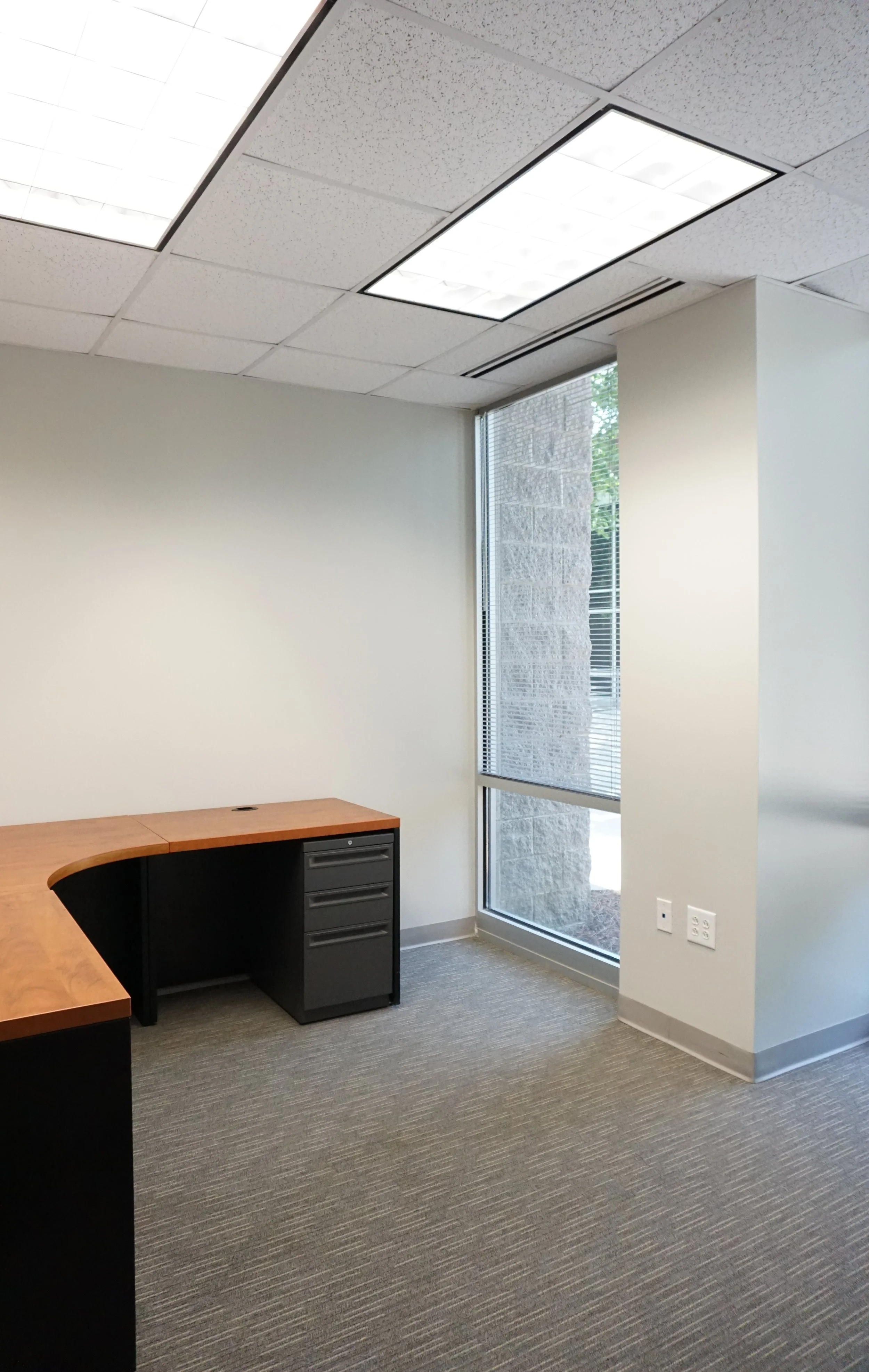 Empty office space with a wooden desk, a window with blinds, and ceiling lights.