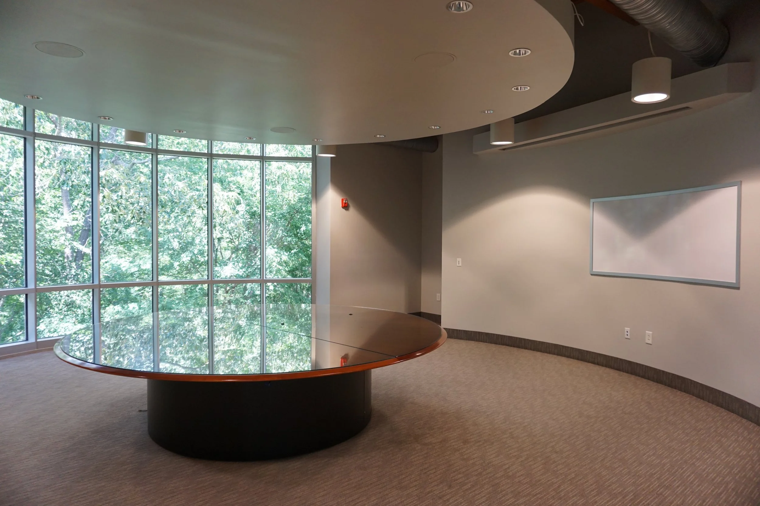 Empty conference room with a curved wall of windows, a large round glass table, and a whiteboard on the wall.