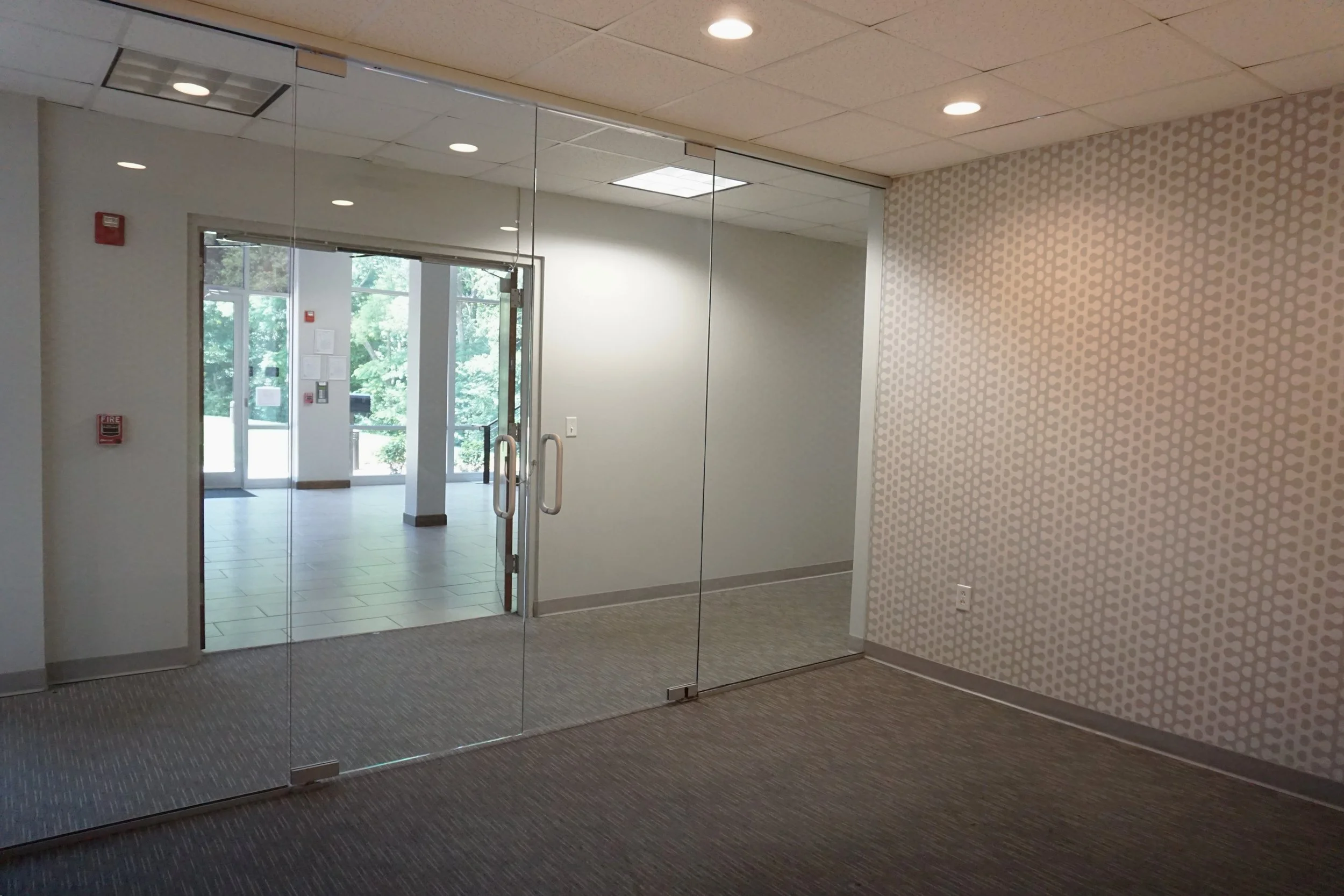 Interior view of a building entrance with glass doors, visible fire alarm and emergency equipment, and large windows showing outside greenery.
