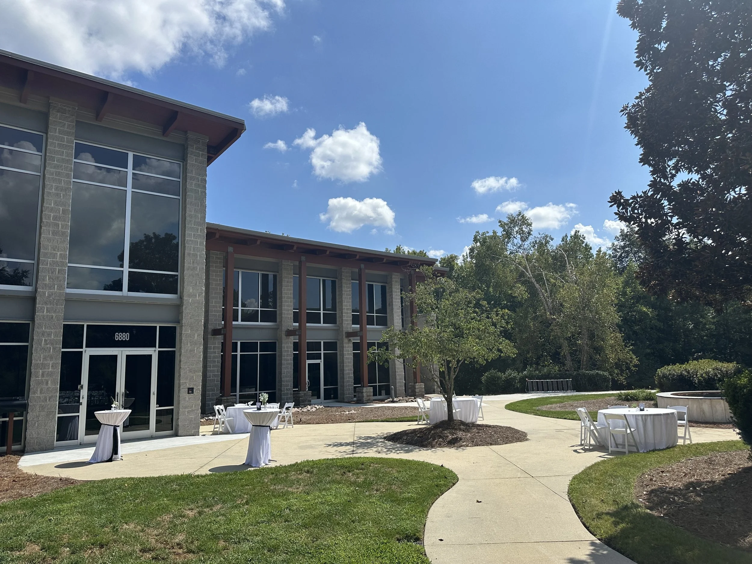 An outdoor event space with white round tables covered with white tablecloths and white chairs, situated on a concrete path in front of a modern building with large glass windows and brick walls, surrounded by grass, trees, and a blue sky with some c