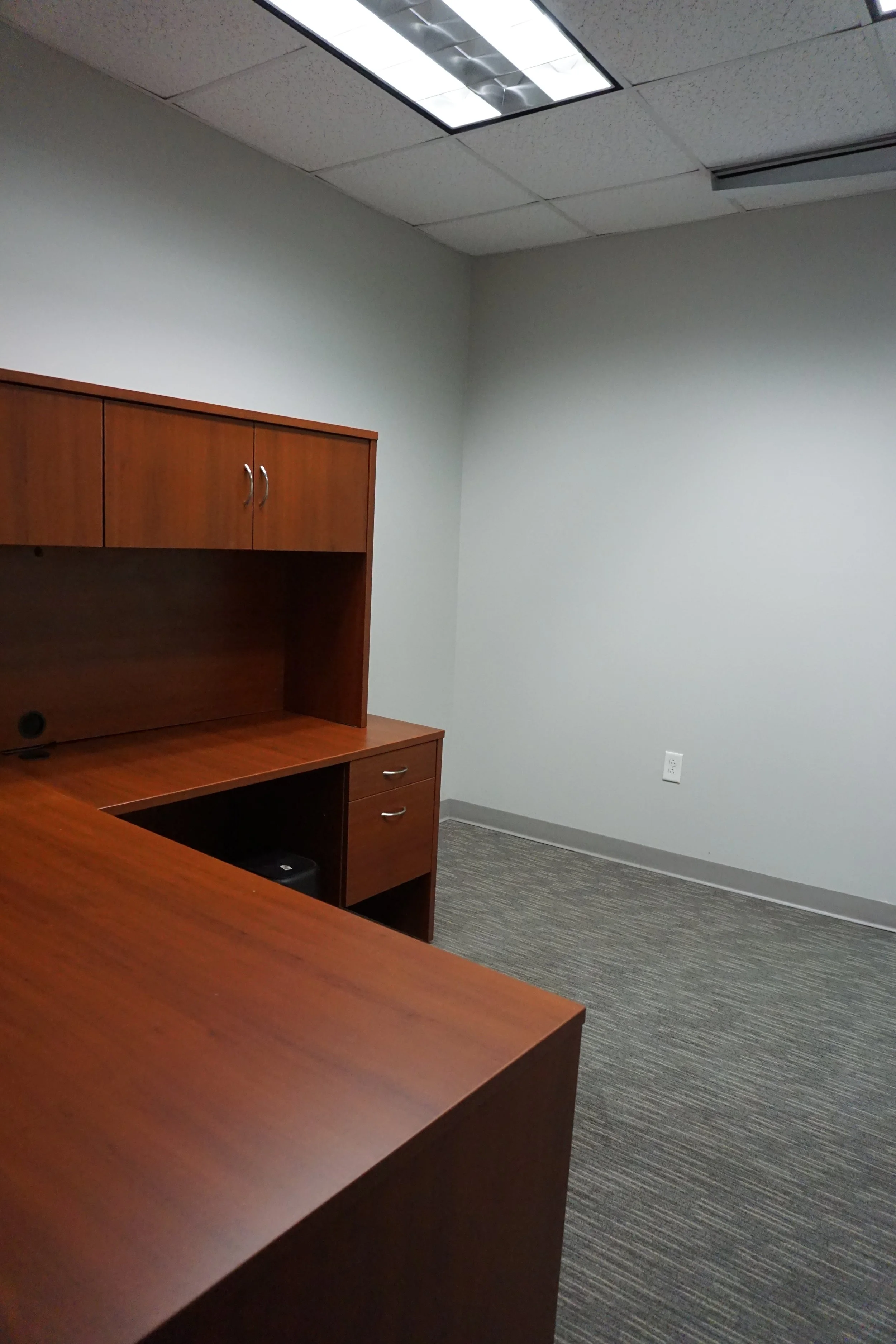 Empty office room with a wooden desk, cabinets, a ceiling light, gray walls, gray carpet, and an electrical outlet.