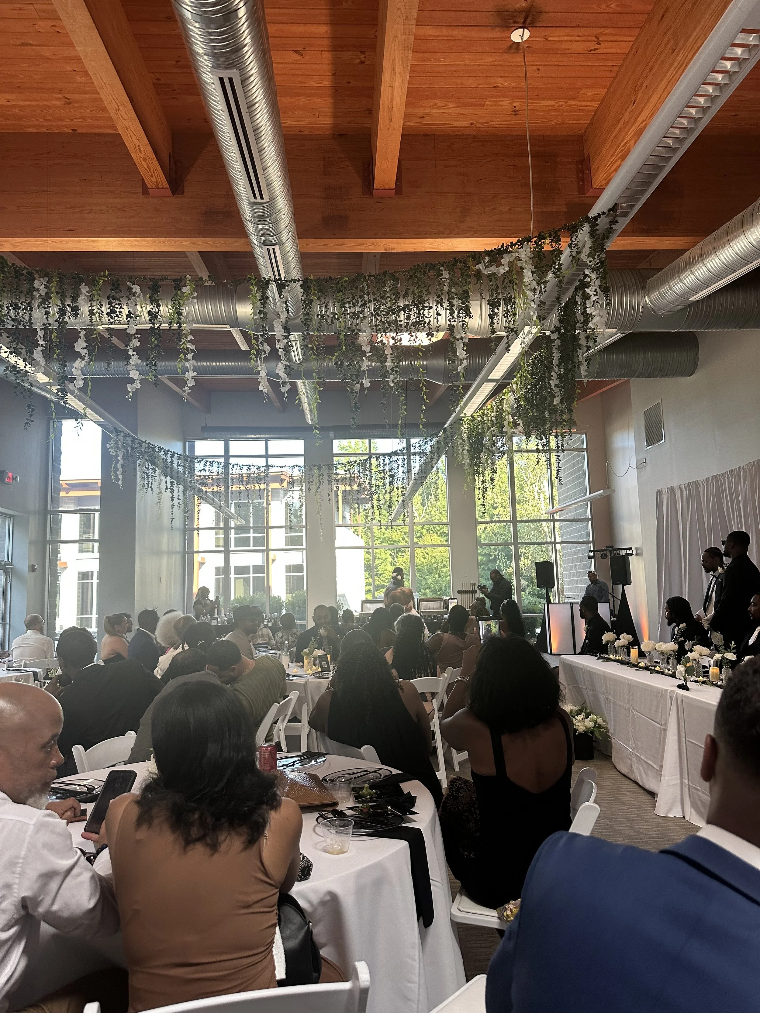 People attending a formal event in a spacious, well-lit room with high ceilings and large windows. Some people are seated at round tables, while others are standing near a long table decorated with white flowers and candles. The room has hanging gree