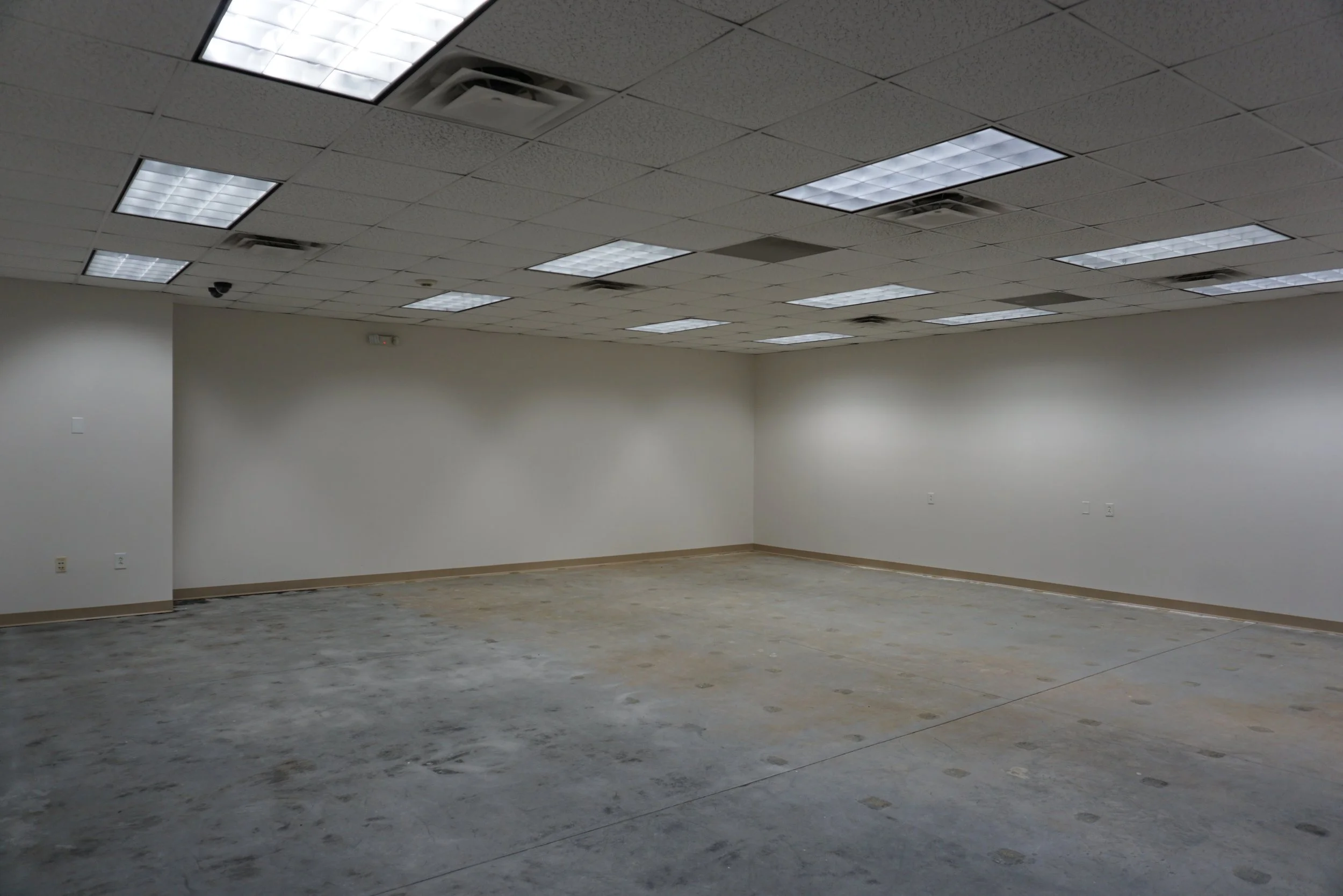 Empty retail or office space with beige walls, fluorescent ceiling lights, and a concrete floor.