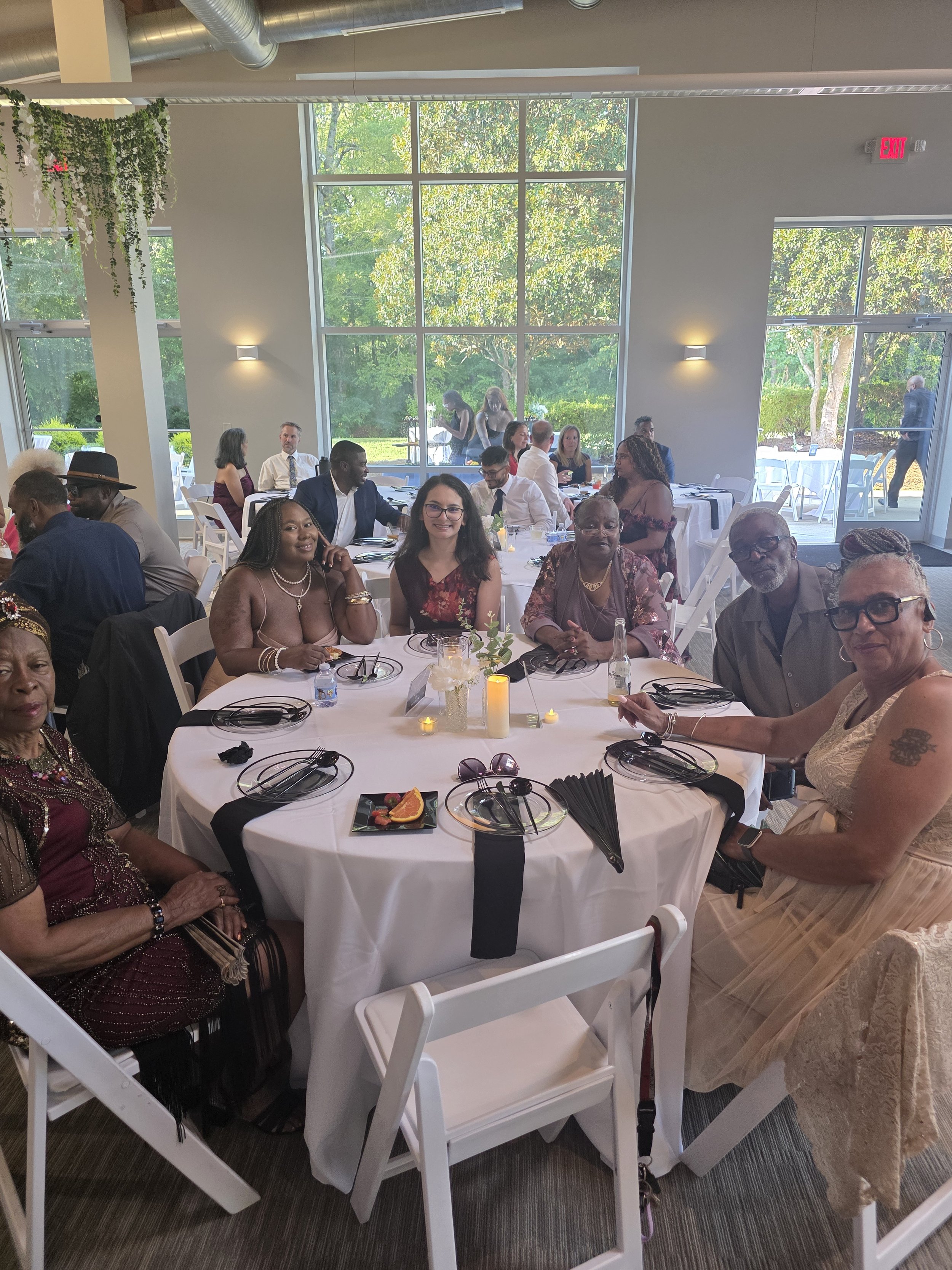 People sitting at a round banquet table with white tablecloths during a daytime event in a bright room with large windows showing greenery outside. The table has black napkins, small candle holders, a floral centerpiece, and some plates with food. So