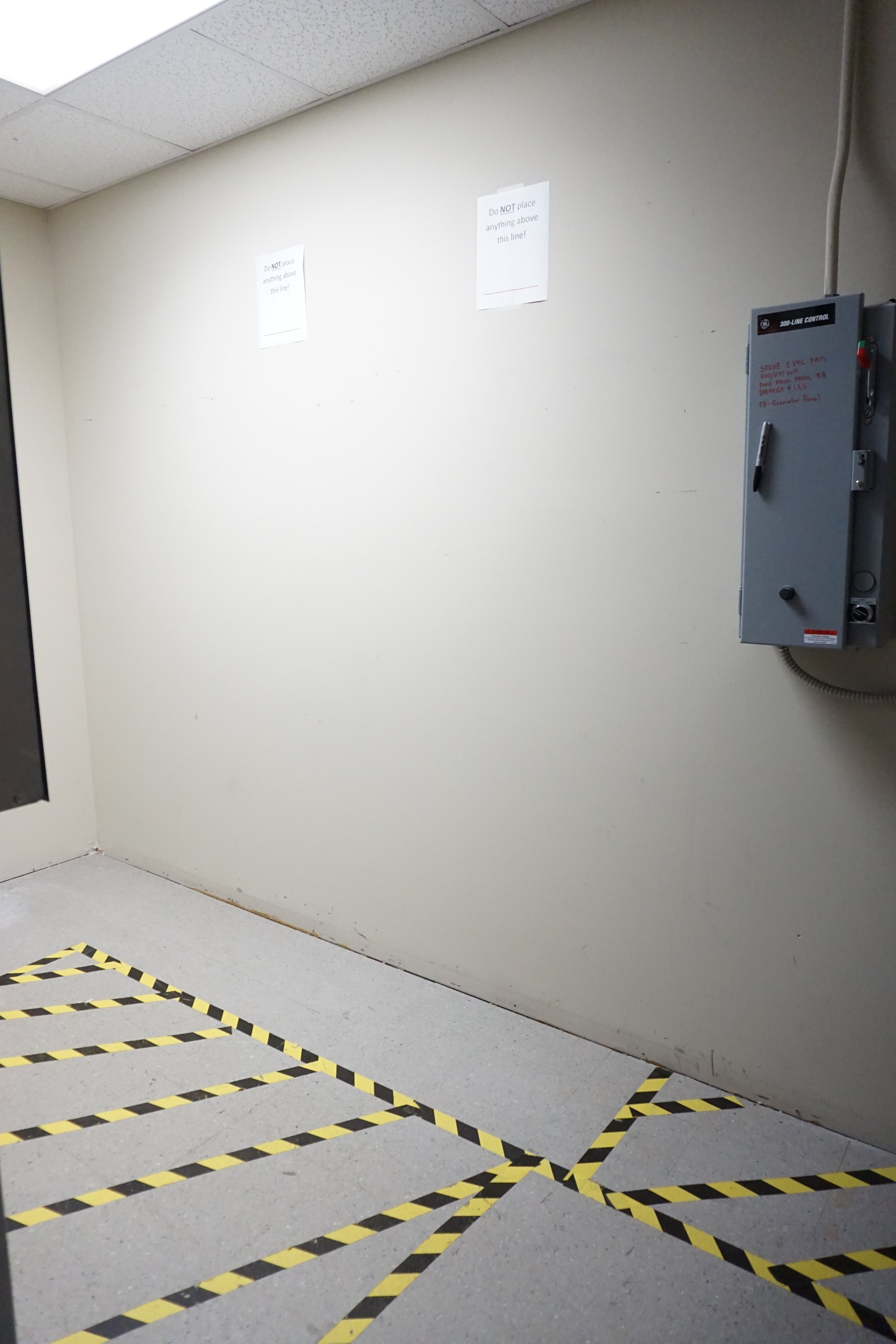 Empty hallway with a white wall, two signs that say 'Do NOT place anything above this line,' a gray electrical box on the right, and a floor marked with black and yellow caution tape in a striped pattern.