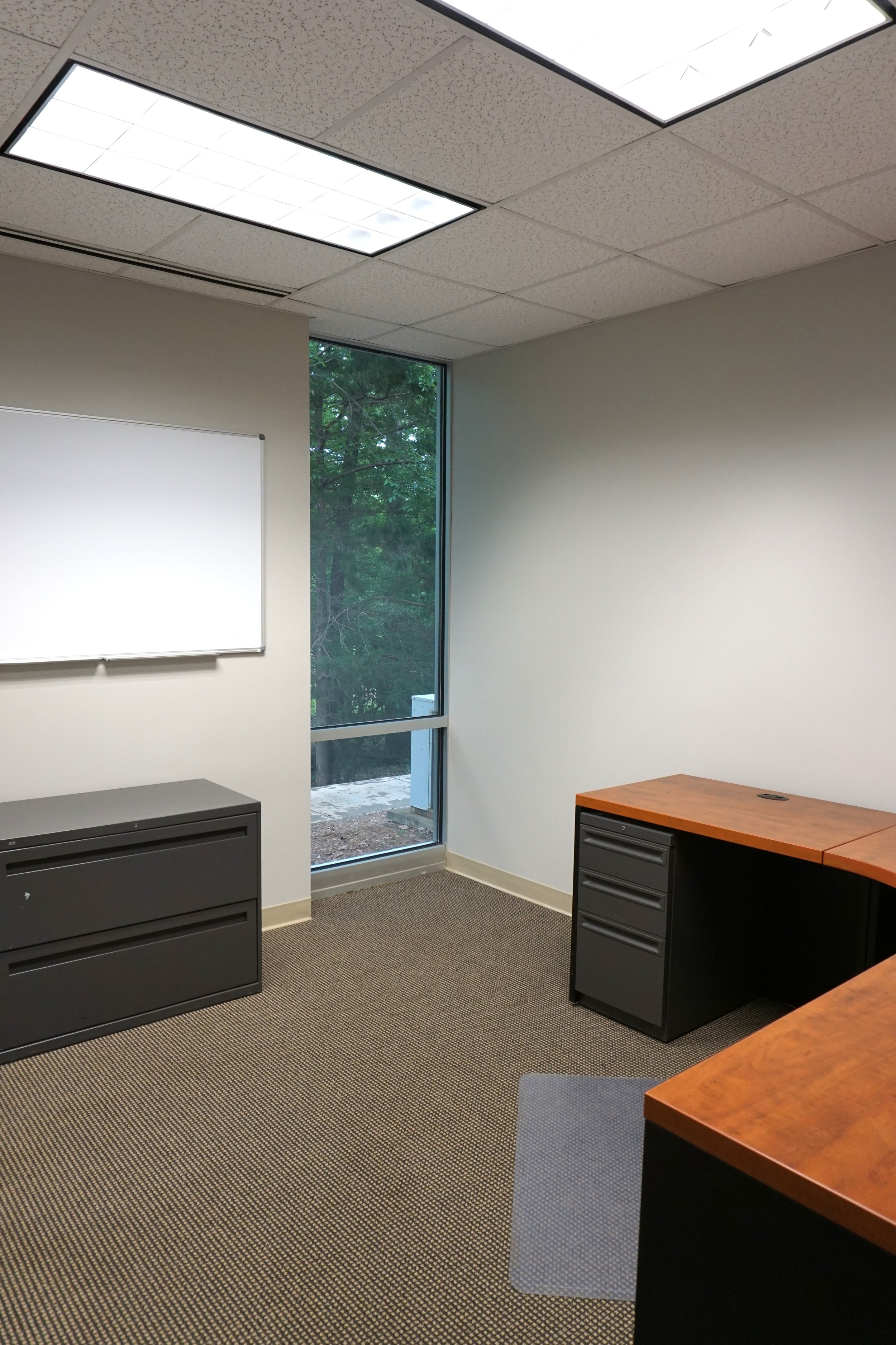 An empty office room with a whiteboard, a window showing trees outside, and two desks with black filing cabinets.