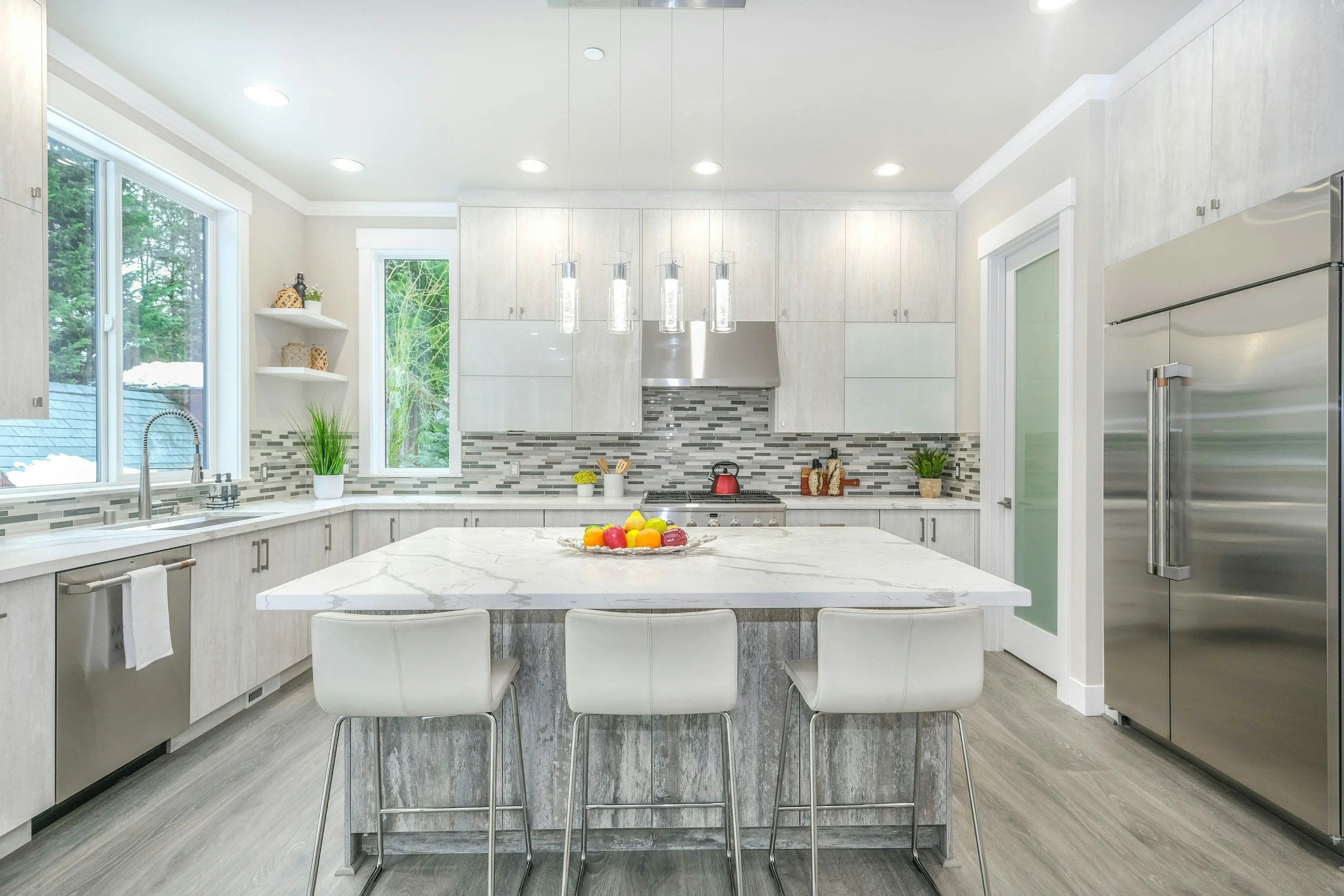 Modern kitchen with white cabinets, gray backsplash, stainless steel appliances, and a large marble island with three white chairs, decorated with a bowl of fruit. Window showing green trees outside.