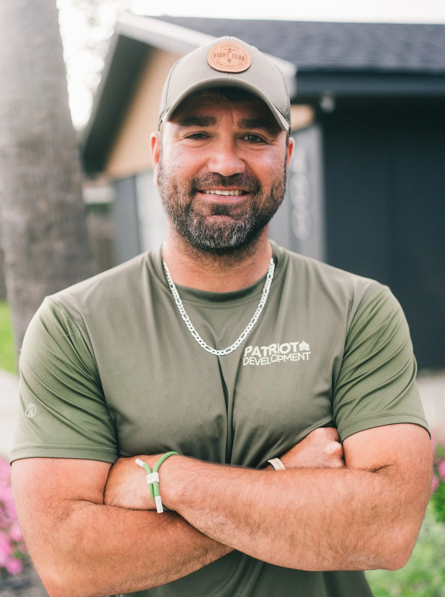A smiling man with a beard, wearing a gray cap, an olive green t-shirt with 'Patriot Development' written on it, a silver chain necklace, and a green wristband, standing outdoors.