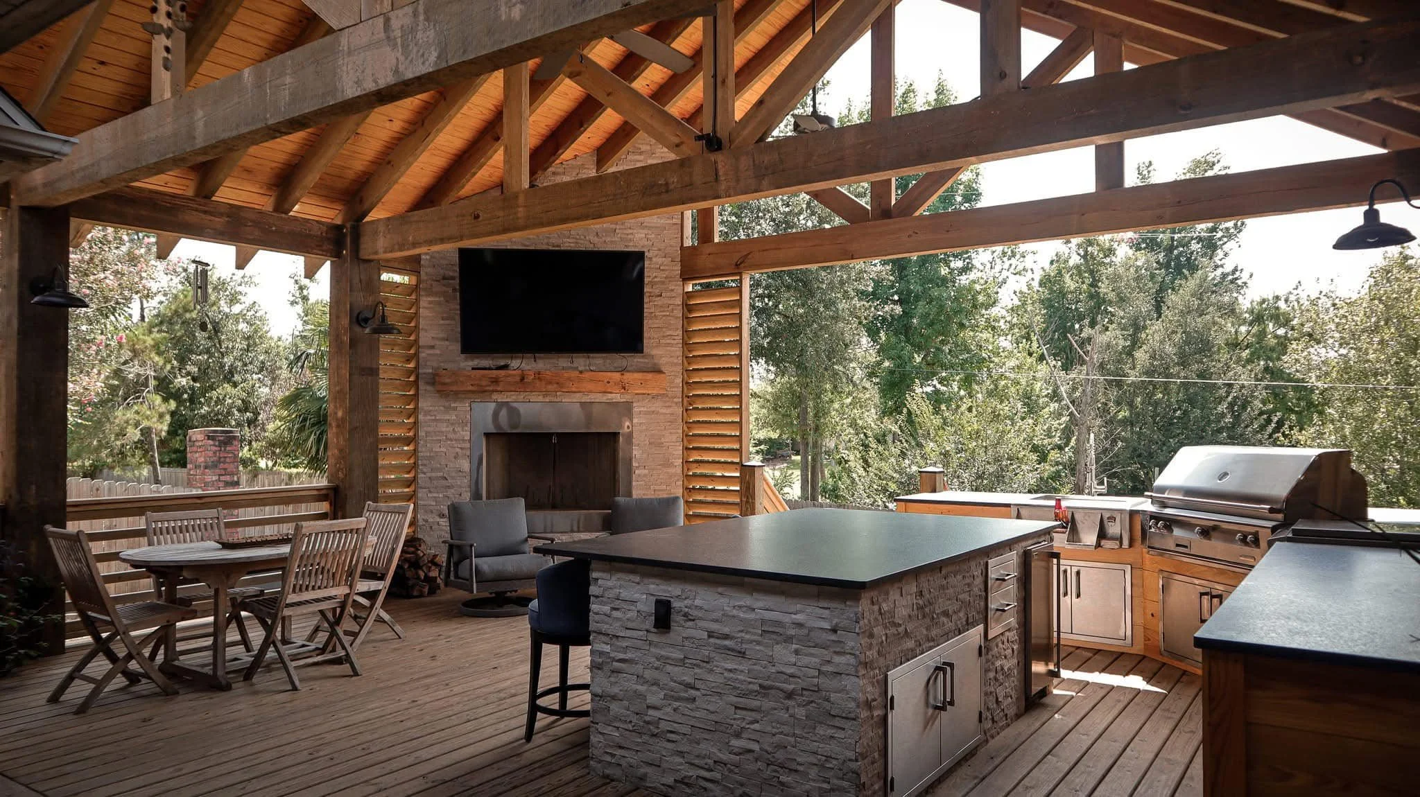 Exterior covered patio with wooden beams, outdoor kitchen with grill, sitting area, dining table with chairs, and a wall-mounted TV, overlooking trees.