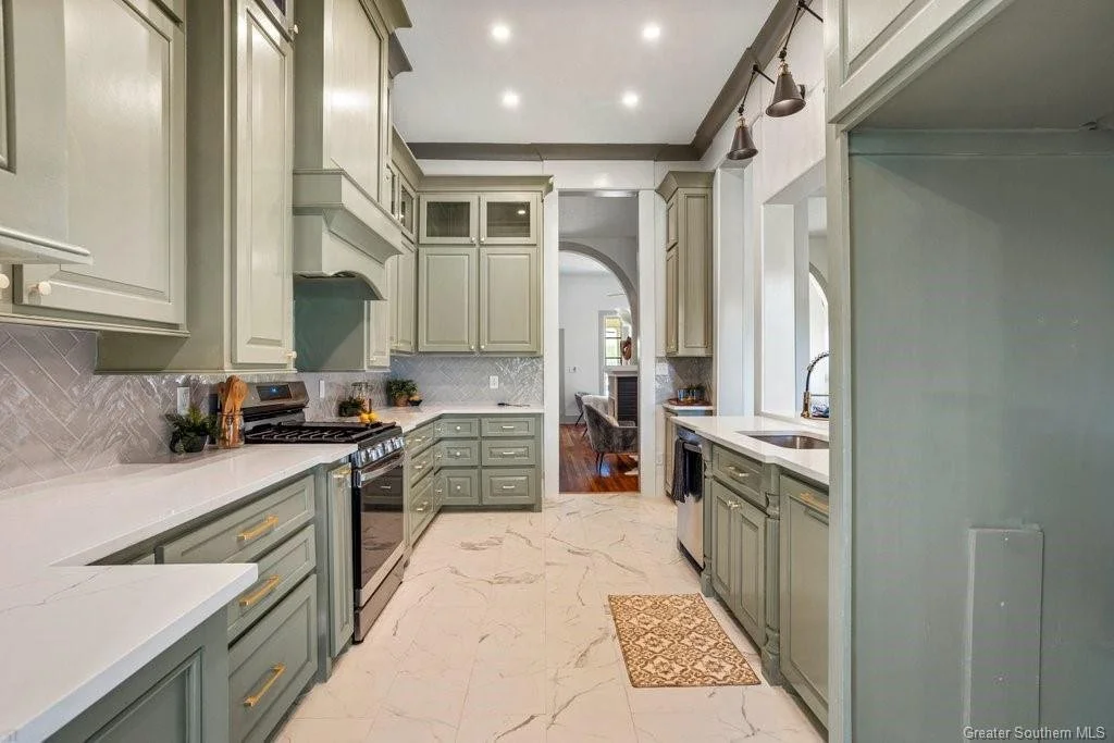 Modern kitchen with light green cabinets, marble countertops, and a marble floor. There is a stove and sink with a window above. A small mat is in front of the sink, and the kitchen opens to a dining and living area with hardwood floors.