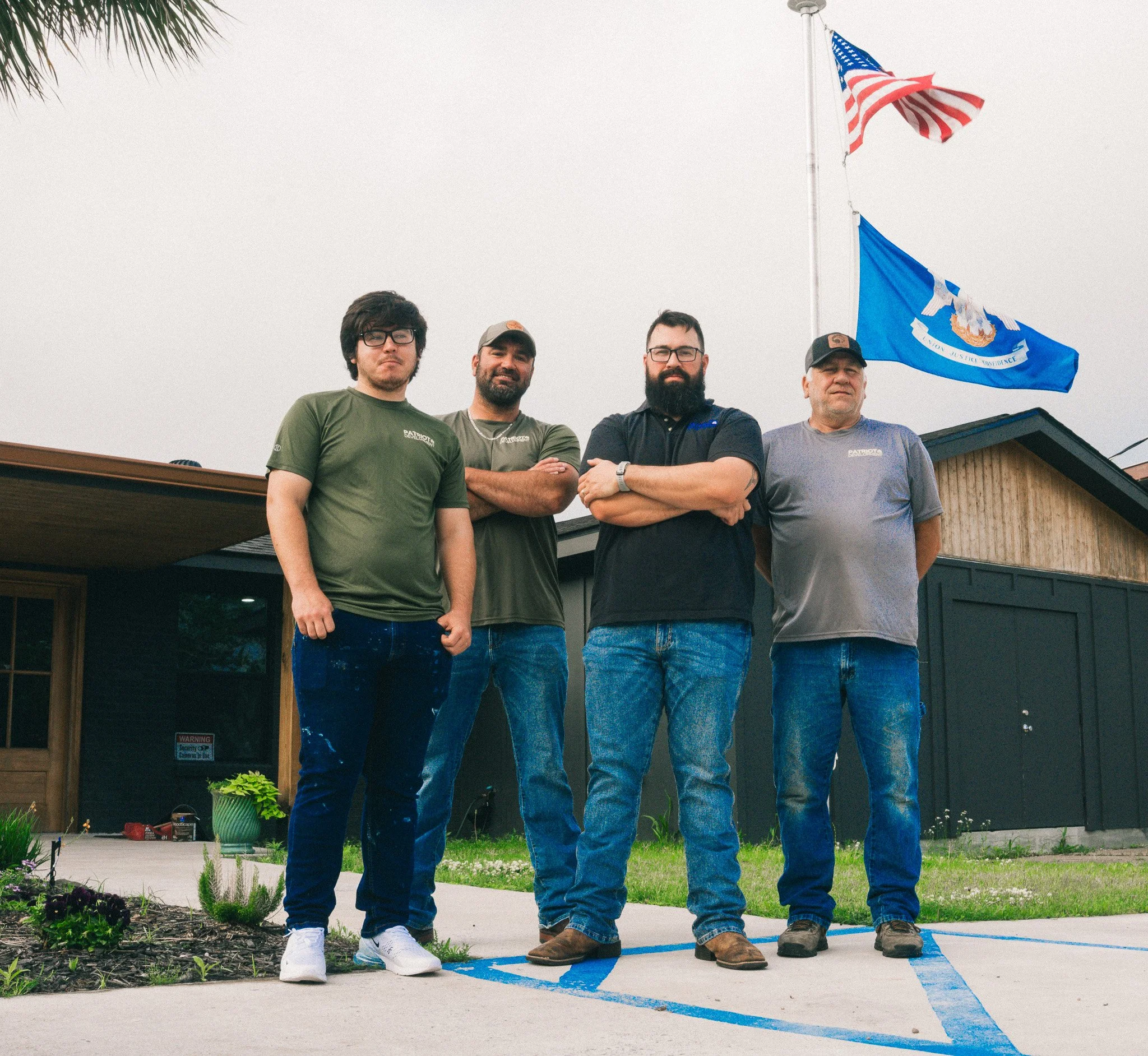 Four men standing outside a building with two American flags against an overcast sky, with a garden and driveway nearby.