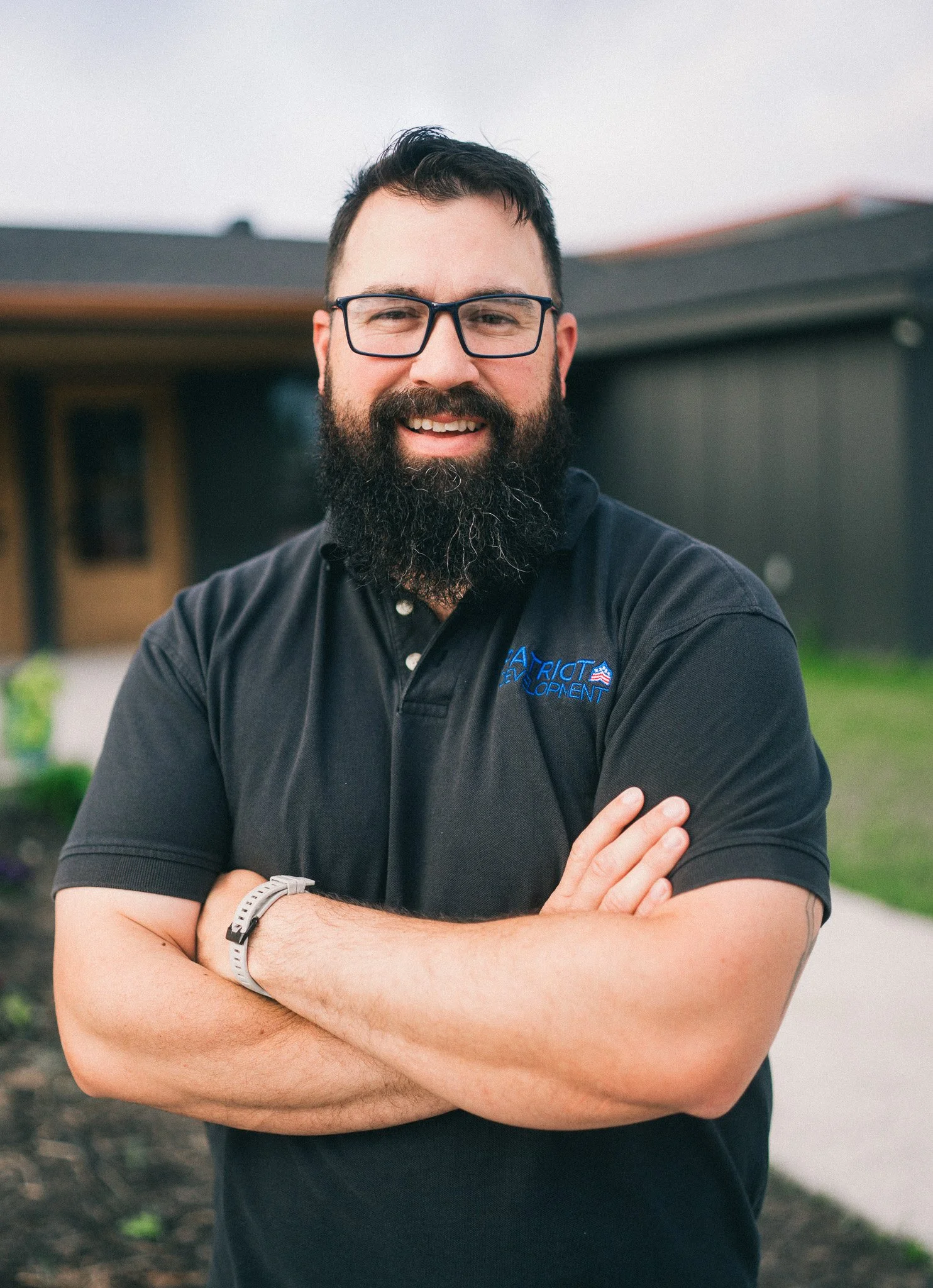 A man with glasses and a beard smiling outdoors with arms crossed, standing in front of a house.