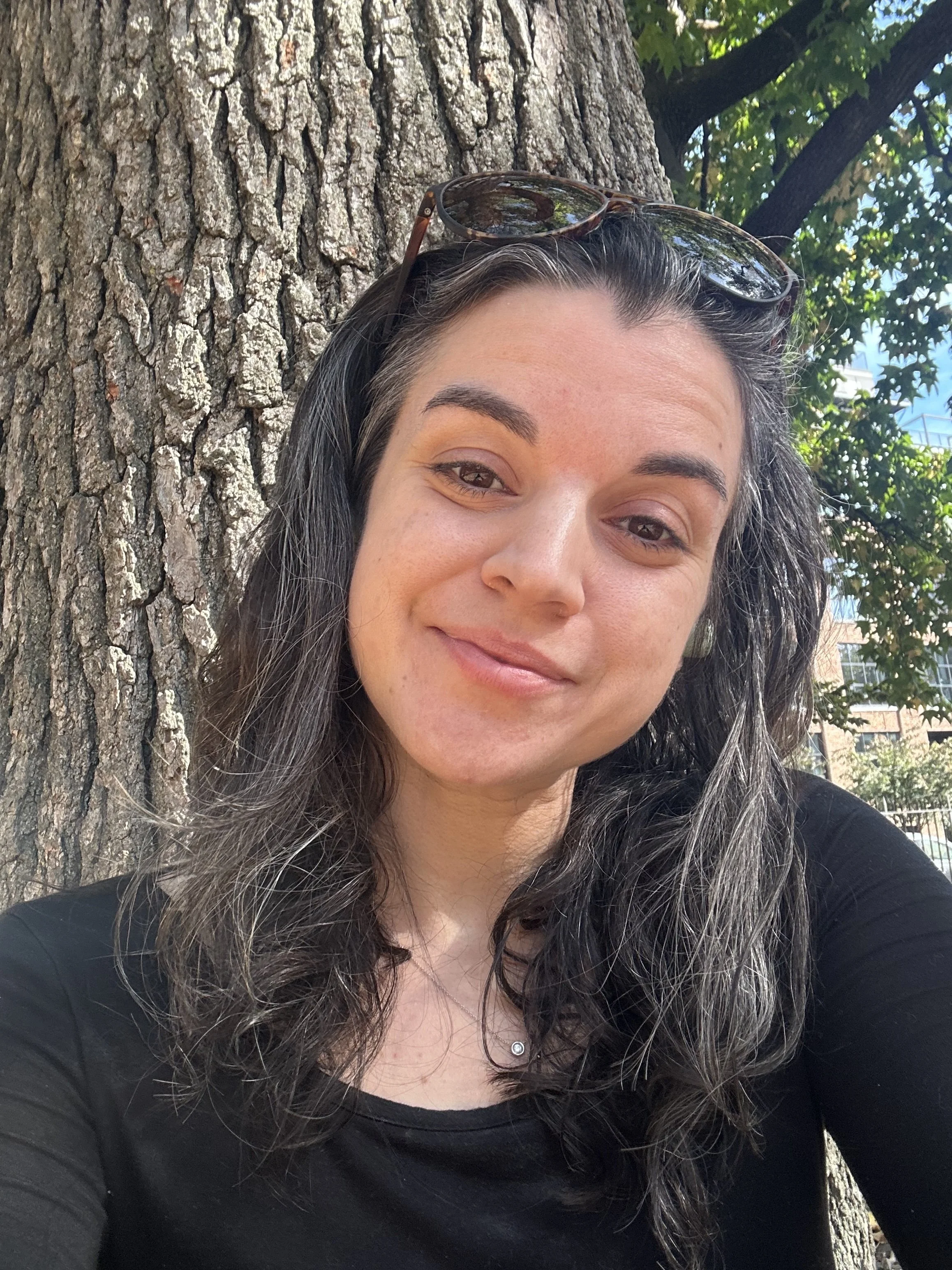 Smiling white woman with dark hair sitting in front of a tree