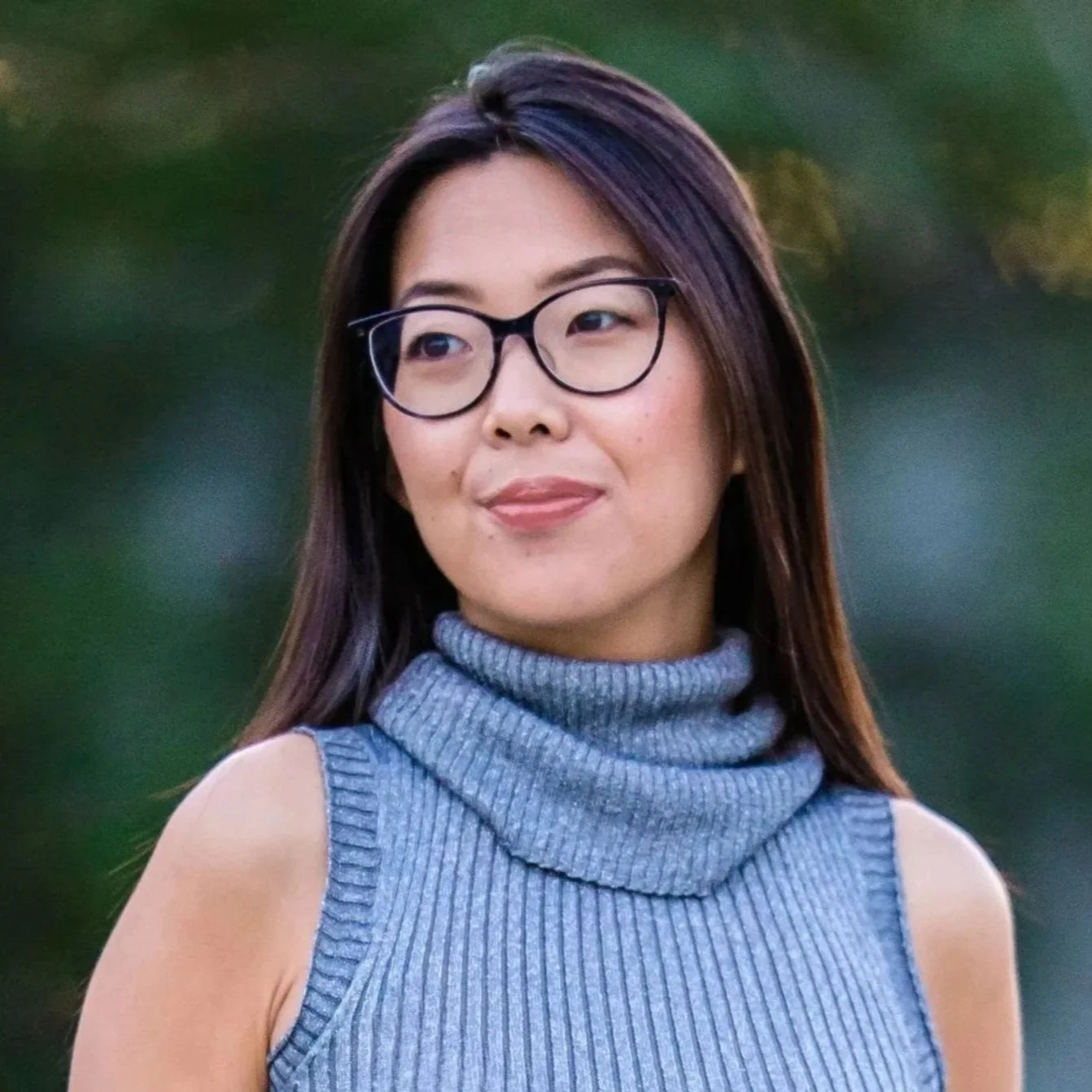 A young woman wearing glasses and a sleeveless gray turtleneck sweater standing outdoors with a blurred green background.