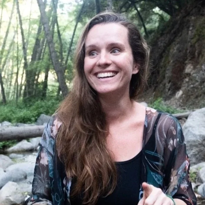 A woman with long brown hair smiling and pointing while sitting outdoors in a forest near rocks and trees.
