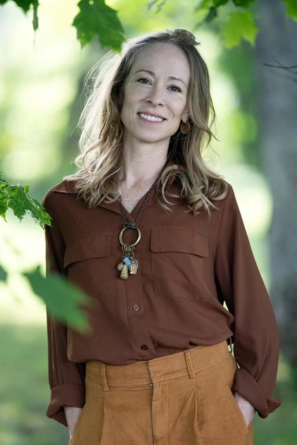 A smiling woman with wavy blonde hair, wearing a brown button-up shirt and tan pants, standing outdoors in a green, leafy background.