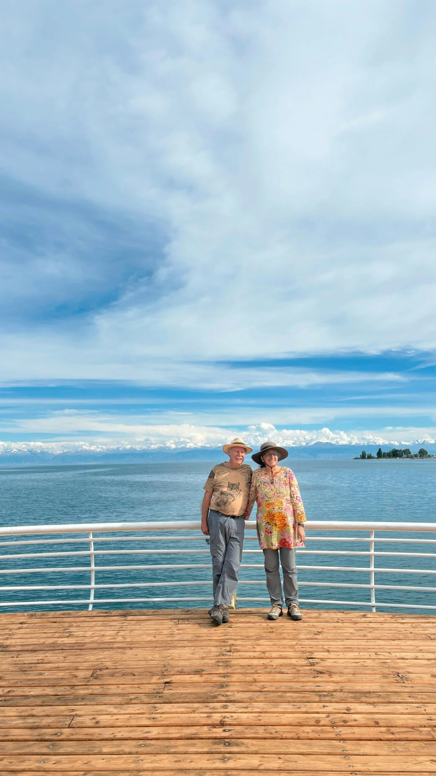 Due persone sorridenti che indossano cappelli, si trovano sulla passerella di una nave affacciata su un lago con montagne innevate sullo sfondo e un cielo nuvoloso